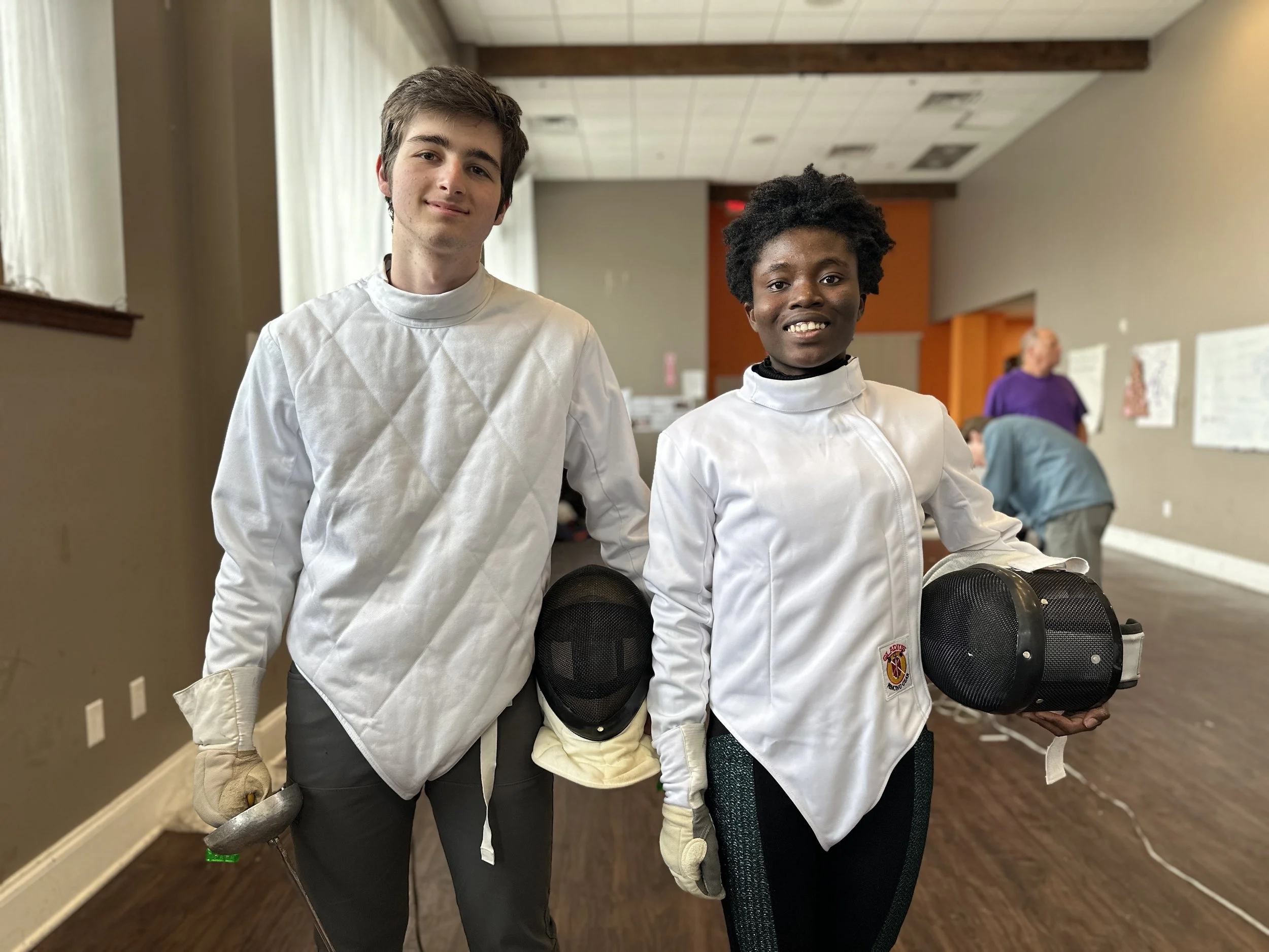 Two young individuals dressed in fencing gear, smiling, holding fencing masks and swords, standing in a room with other people in the background.