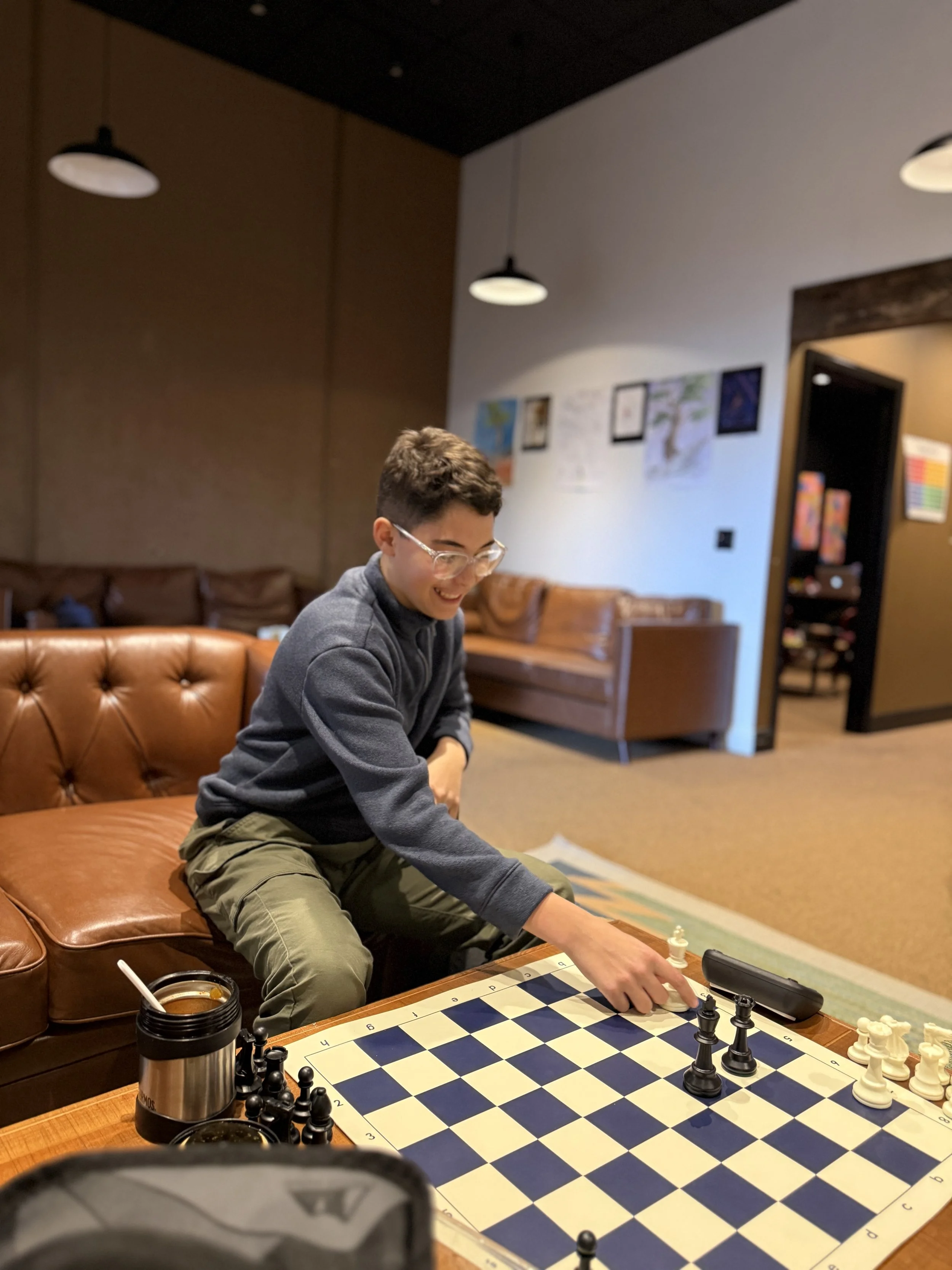 Students sitting at a long table in a classroom, taking notes, with a teacher writing on a whiteboard in the background.