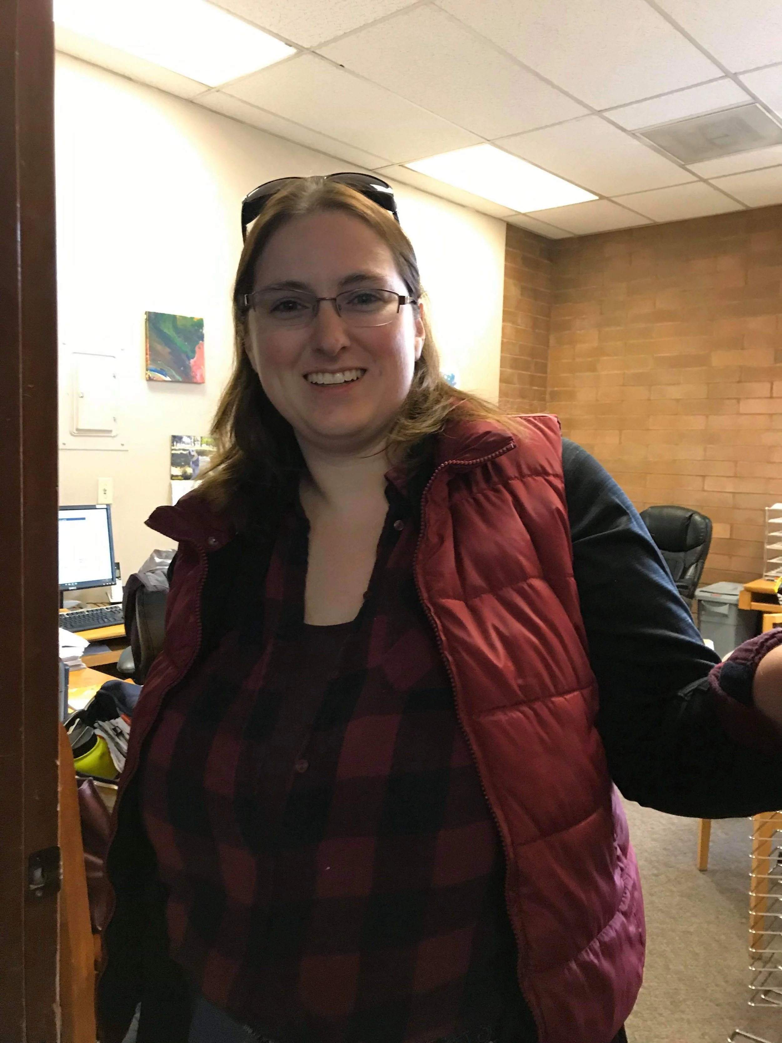 A woman with glasses smiling, wearing a red vest over a plaid shirt, standing in an office.