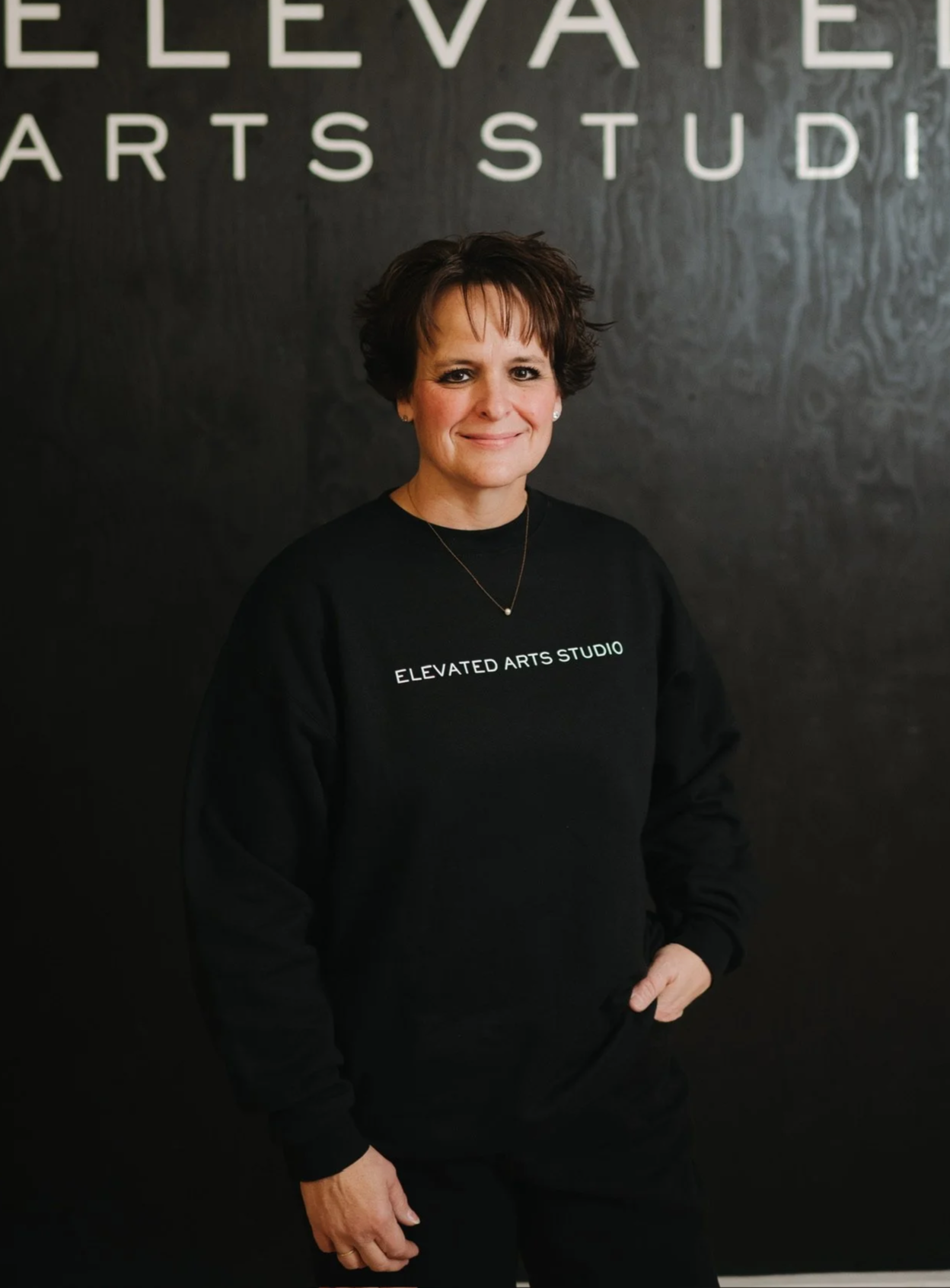 A woman with short dark hair wearing a black sweatshirt with white text that says 'ELEVATED ARTS STUDIO,' standing in front of a dark wall with large white letters spelling 'ELEVATED ARTS STUDIO.'