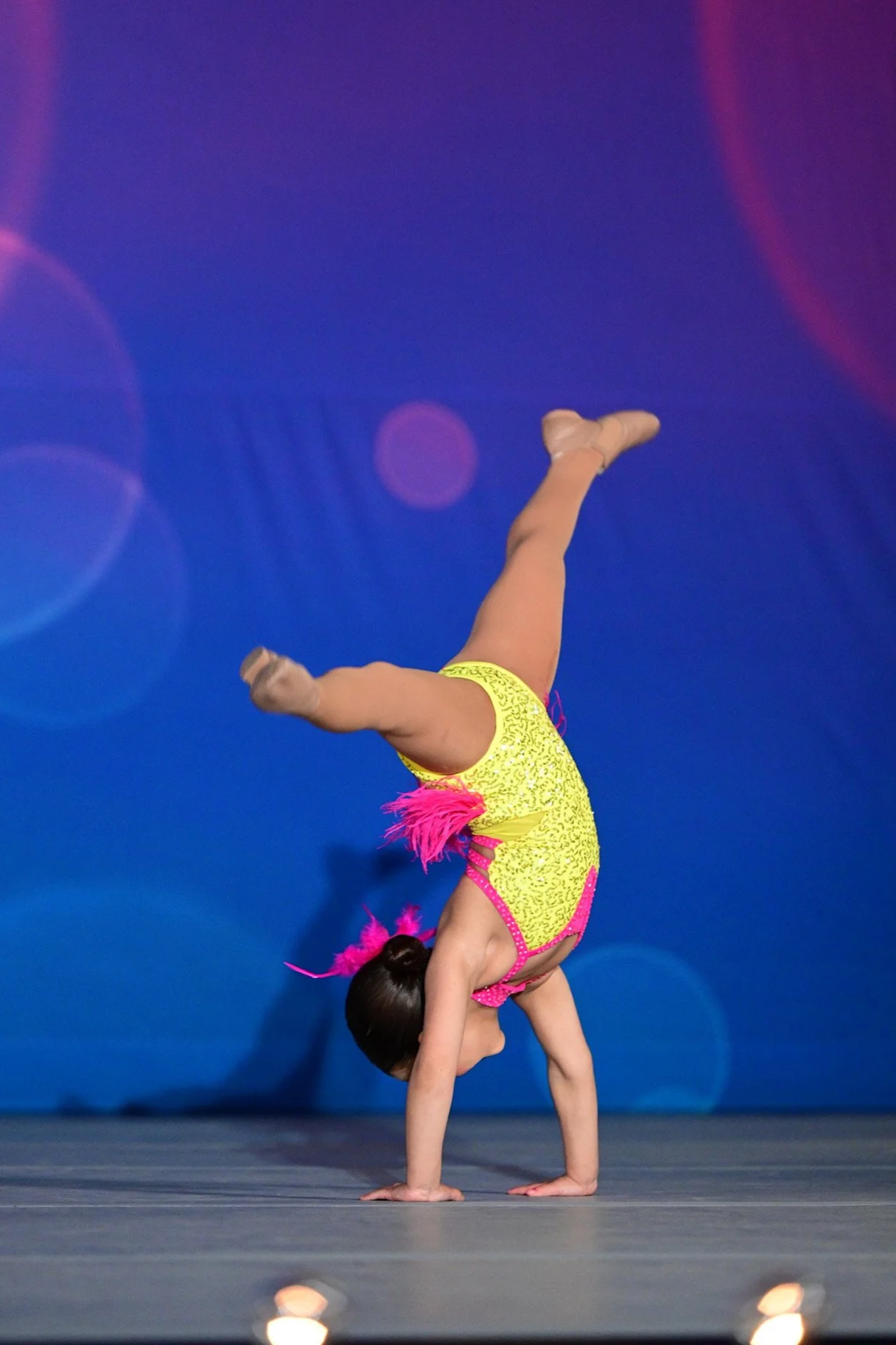A young girl performing a handstand on stage, wearing a yellow and pink gymnastics leotard with pink feather decorations and a pink feathered hair accessory, against a blue background with lights.