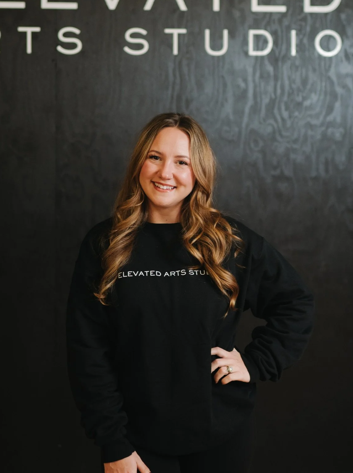 A young woman with long, wavy blonde hair smiling while standing in front of a black wall with white text partially visible behind her. She is wearing a black sweatshirt that reads 'ELEVATED ARTS STUDIO'.