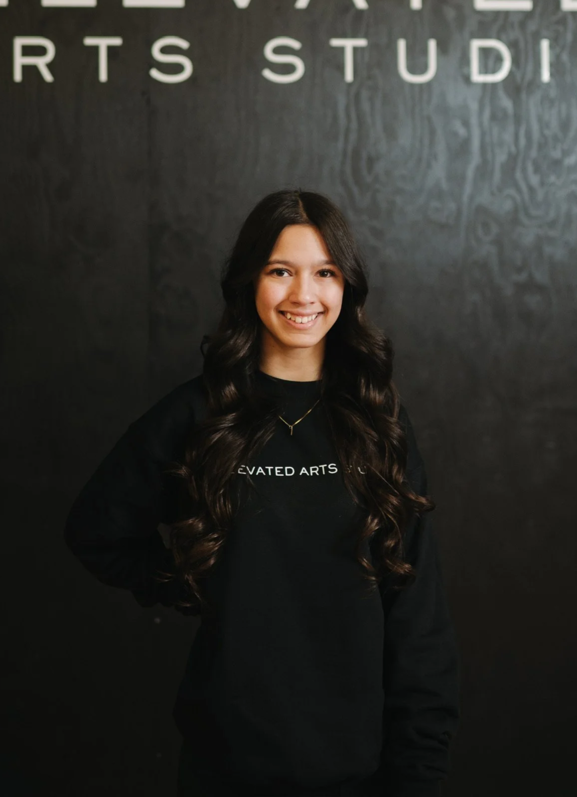 A young woman with long, dark wavy hair smiling, wearing a black sweatshirt with white text and a necklace, standing in front of a black textured wall with partial white letters.