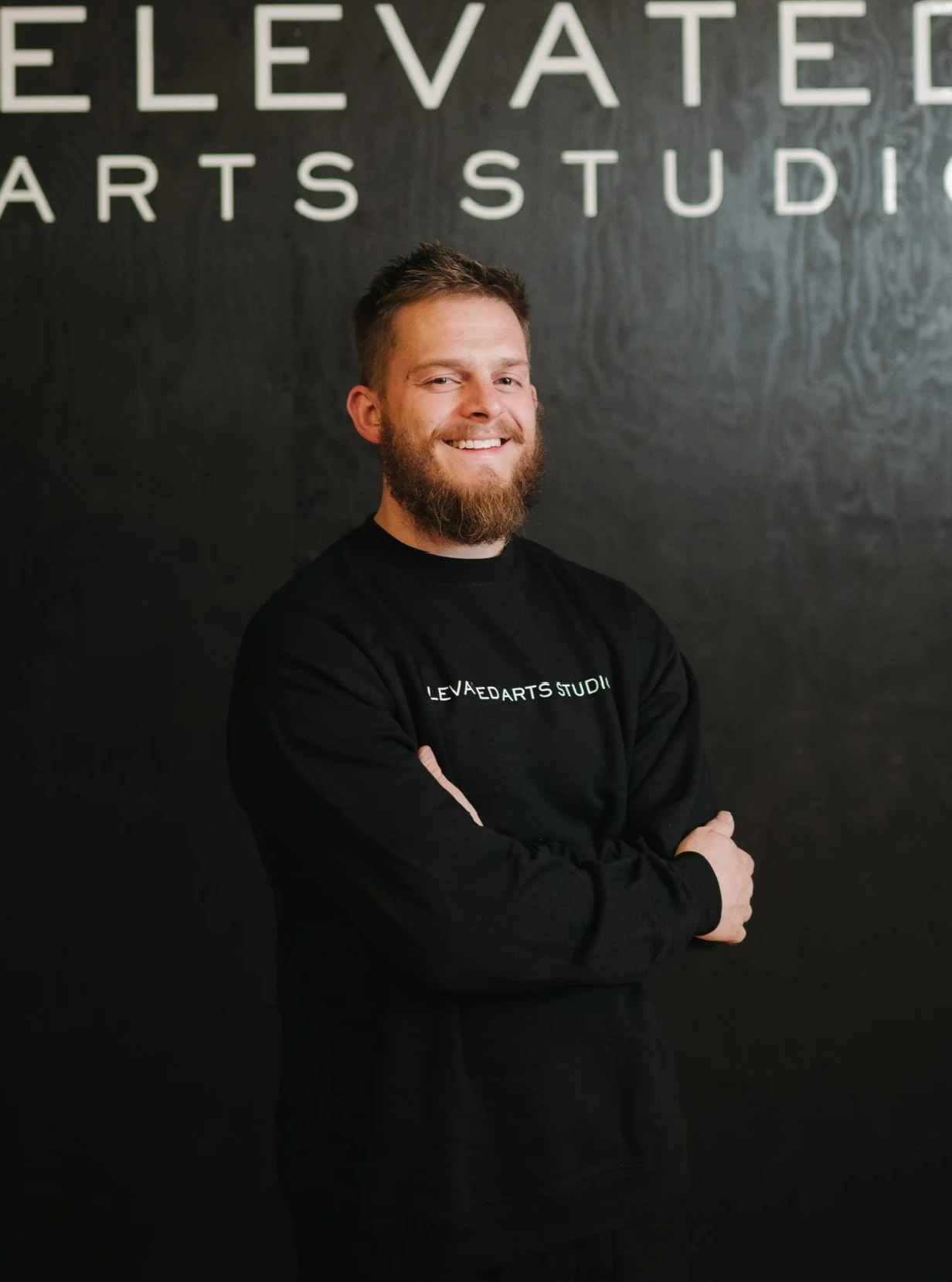 A smiling man with a beard and short hair standing with arms crossed in front of a black wall with white lettering that reads 'ELEVATED ARTS STUDIO'. He is wearing a black sweatshirt with similar text.