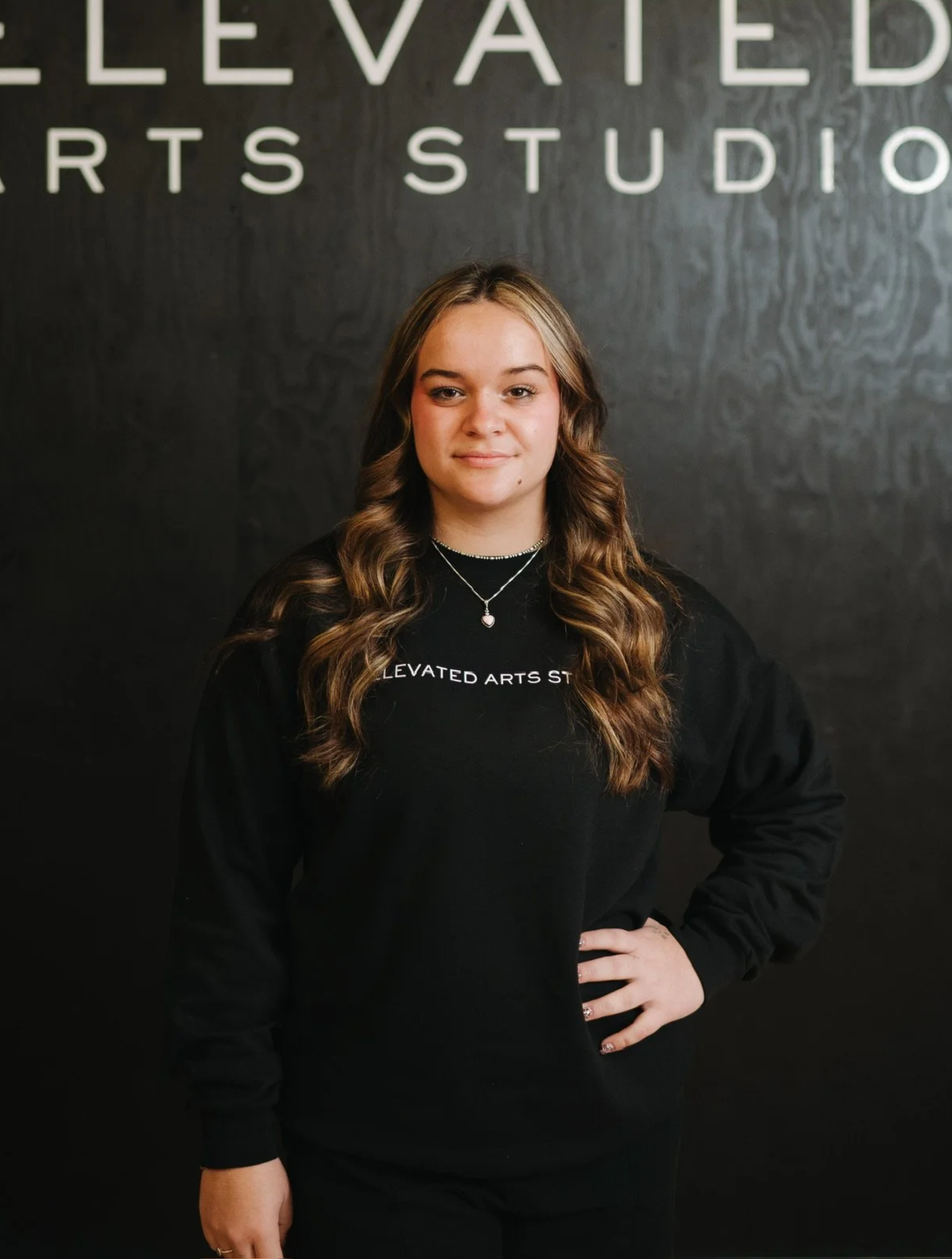 A young woman with long wavy brown hair standing in front of a black wall with white letters that say 'Elevated Arts Studio.' She is wearing a black sweatshirt with the same words and pearl necklaces, with her left hand on her hip.