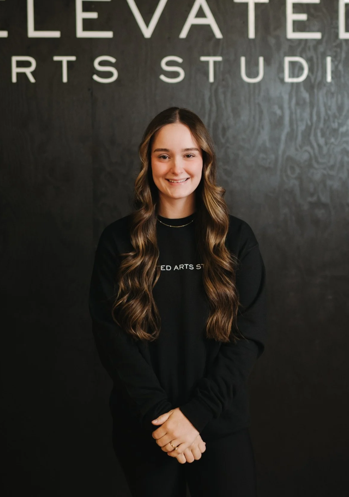 A young woman with long wavy brown hair standing in front of a dark wall with large white letters that spell ''ELEVATED ARTS STUDIO''. She is smiling, wearing a black sweatshirt with the same text and has her hands clasped in front of her.