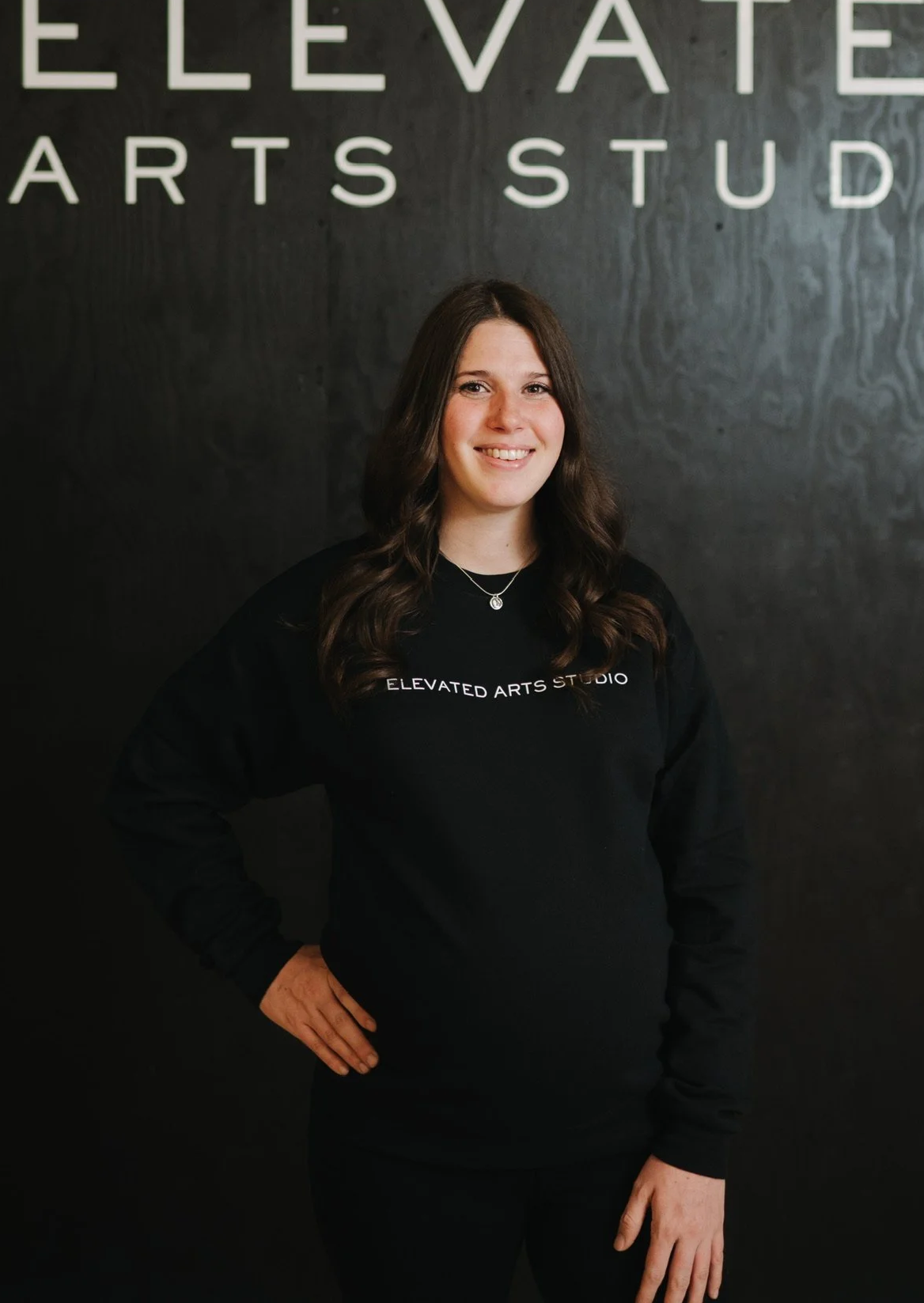 A woman with long, wavy brown hair standing in front of a black wall with the words 'ELEVATED ARTS STUDIO' written on it. She is wearing a black sweatshirt with 'ELEVATED ARTS STUDIO' printed on it in white, and smiling with one hand on her hip.