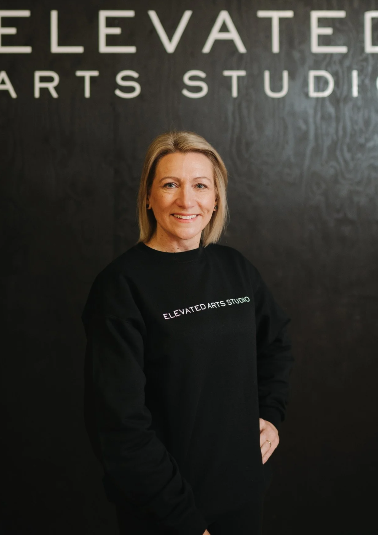 Smiling woman with shoulder-length blonde hair wearing a black sweatshirt that says 'ELEVATED ARTS STUDIO' standing against a black wall with white lettering spelling 'ELEVATED ARTS STUDIO'.