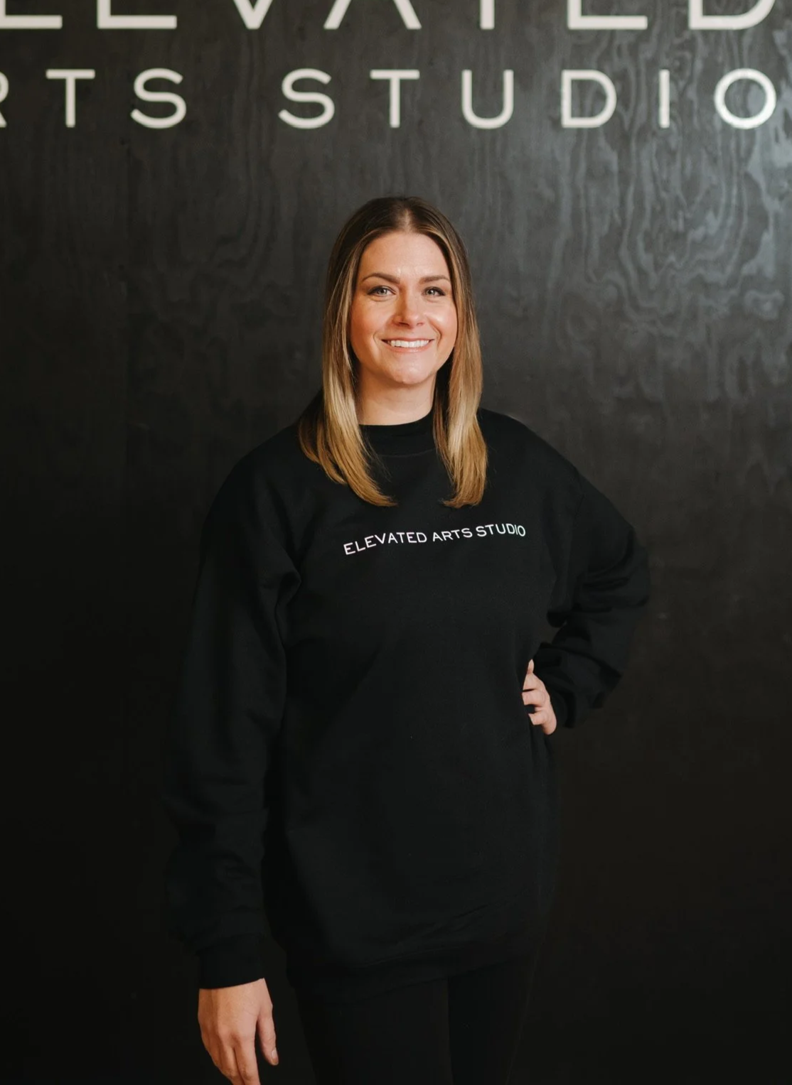 A woman with shoulder-length hair, smiling, stands in front of a dark wall with white letters spelling "ELEVATED ARTS STUDIO." She wears a black sweatshirt with the same text printed on it.