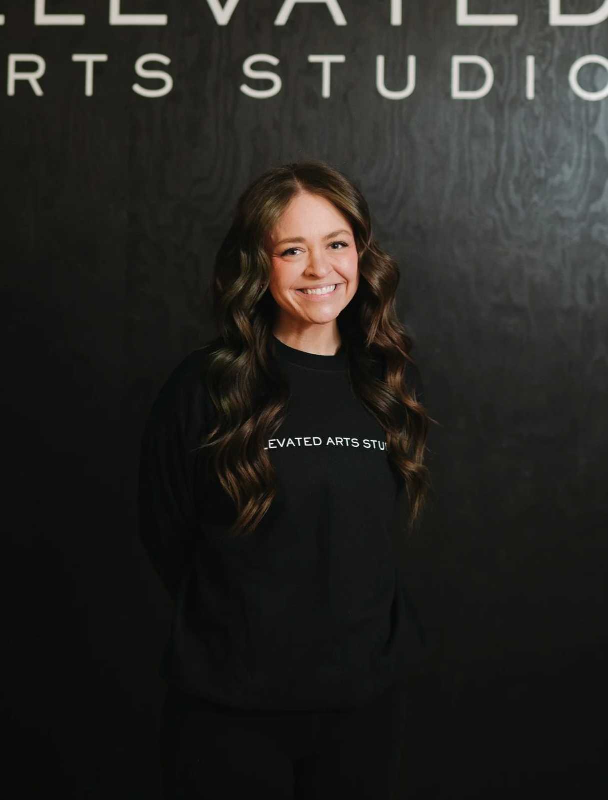 A woman with long, wavy brown hair smiling, wearing a black sweatshirt with white text, standing in front of a black wall with white letters that spell 'ELEVATED ARTS STUDIO'.