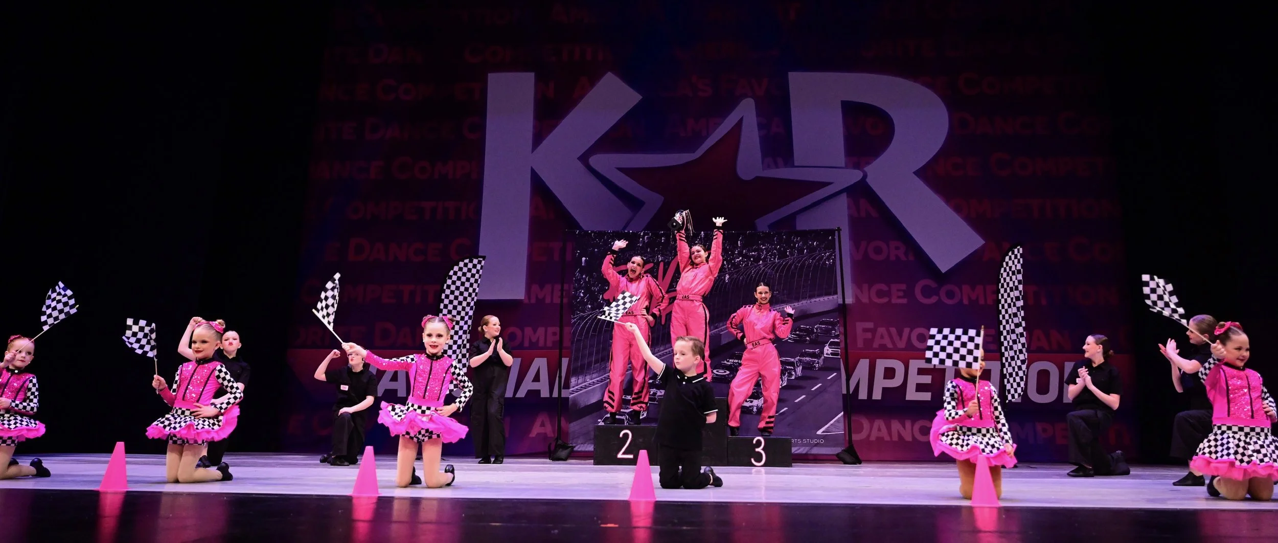 Kids' dance competition on stage with children in pink and black costumes, judges, checkered flags, winner on the podium, and a large 'KAR' logo in the background.