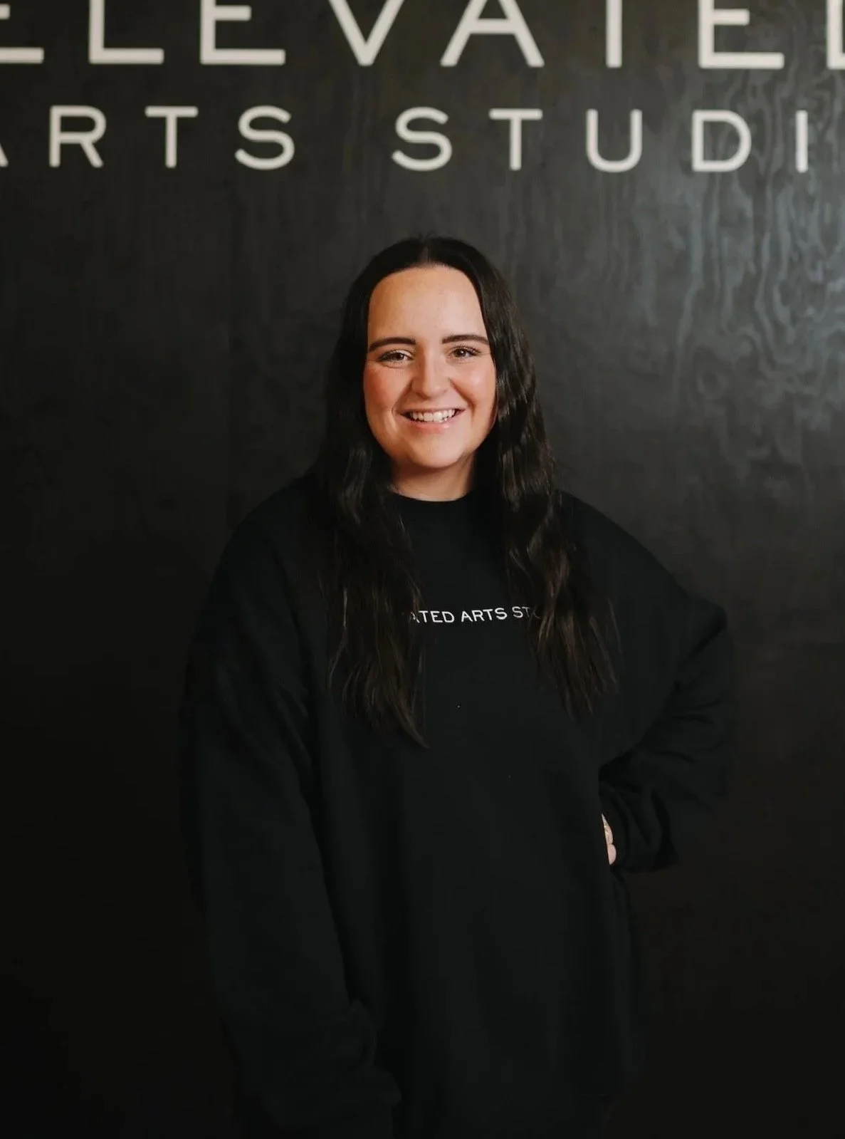 A smiling woman with long dark hair standing in front of a dark wall with the words 'Elevated Arts Studio' partially visible above her head. She is wearing a black sweatshirt with the studio's name on it.