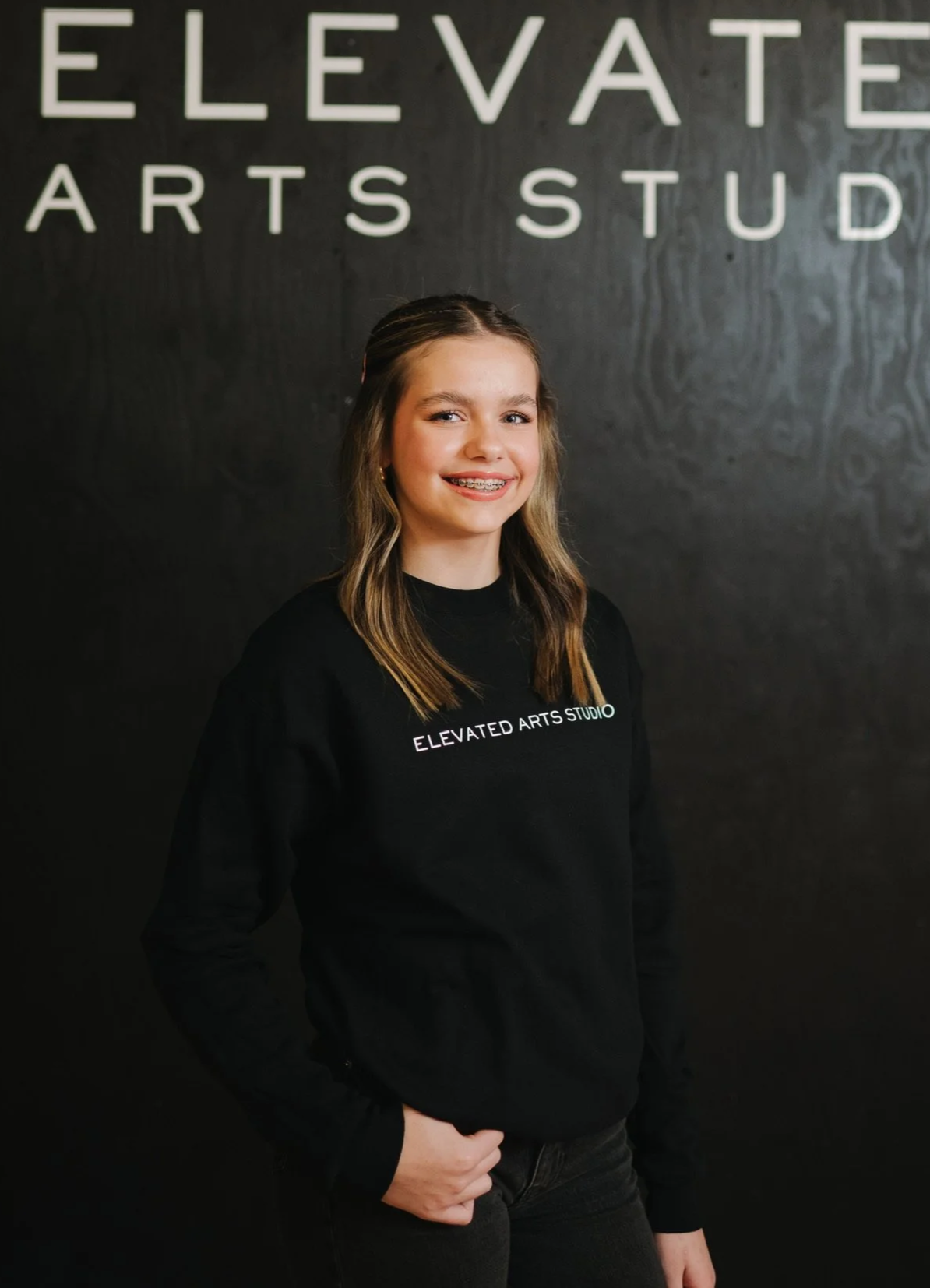 A young girl with light brown hair wearing braces and a black sweatshirt with white text that reads "ELEVATED ARTS STUDIO" stands in front of a black wall with large white letters spelling "ELEVATED ARTS STUDIO."