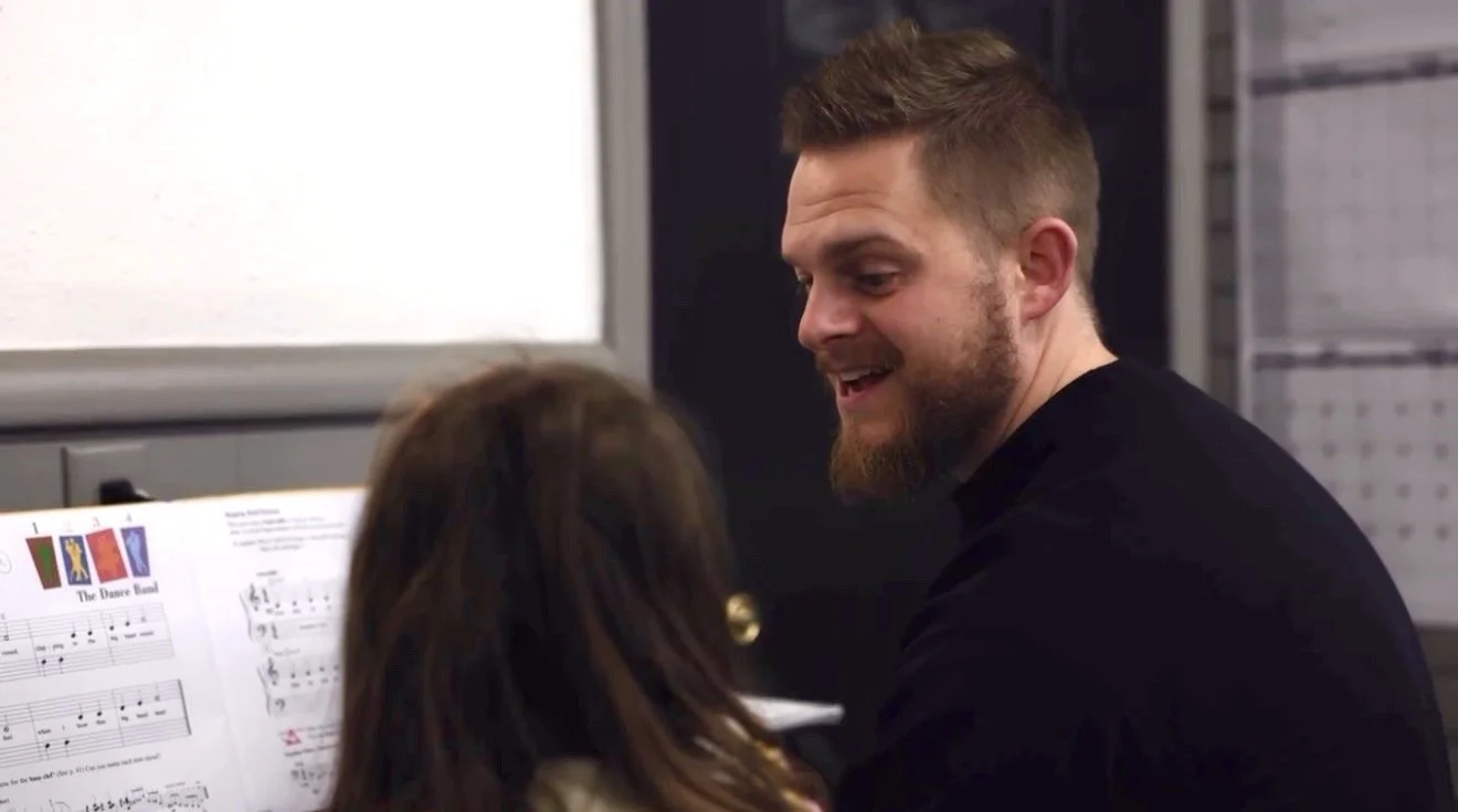 A man with a beard and short brown hair smiling and talking to a girl with long brown hair, who is facing away from the camera, in front of sheet music.