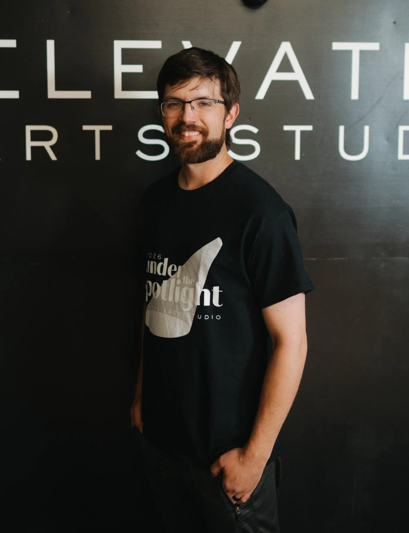 A man with glasses and a beard standing in front of a black wall with white letters spelling 'ELEVATED ARST STUDIO', smiling and wearing a black t-shirt and black pants.