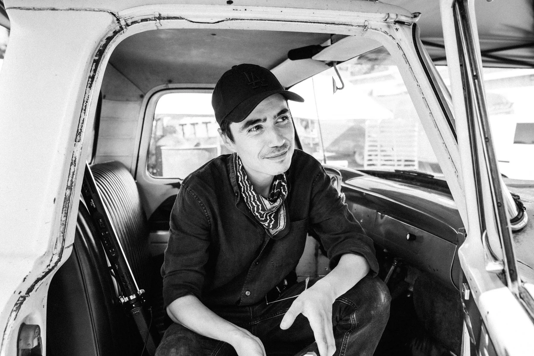 Antonio Diaz sitting inside a vintage vehicle, smiling and looking to the side. He is wearing a baseball cap, a bandana around his neck, and a dark shirt.