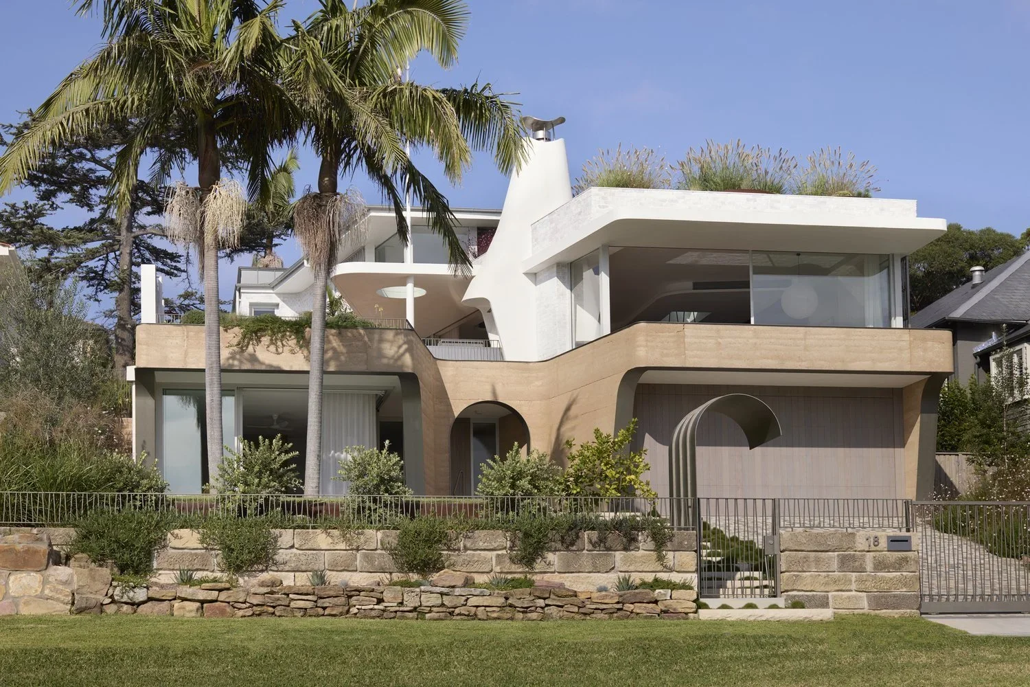 Modern multi-story house with white and beige exterior, surrounded by greenery and palm trees, with a landscaped front yard and steps leading to the entrance.
