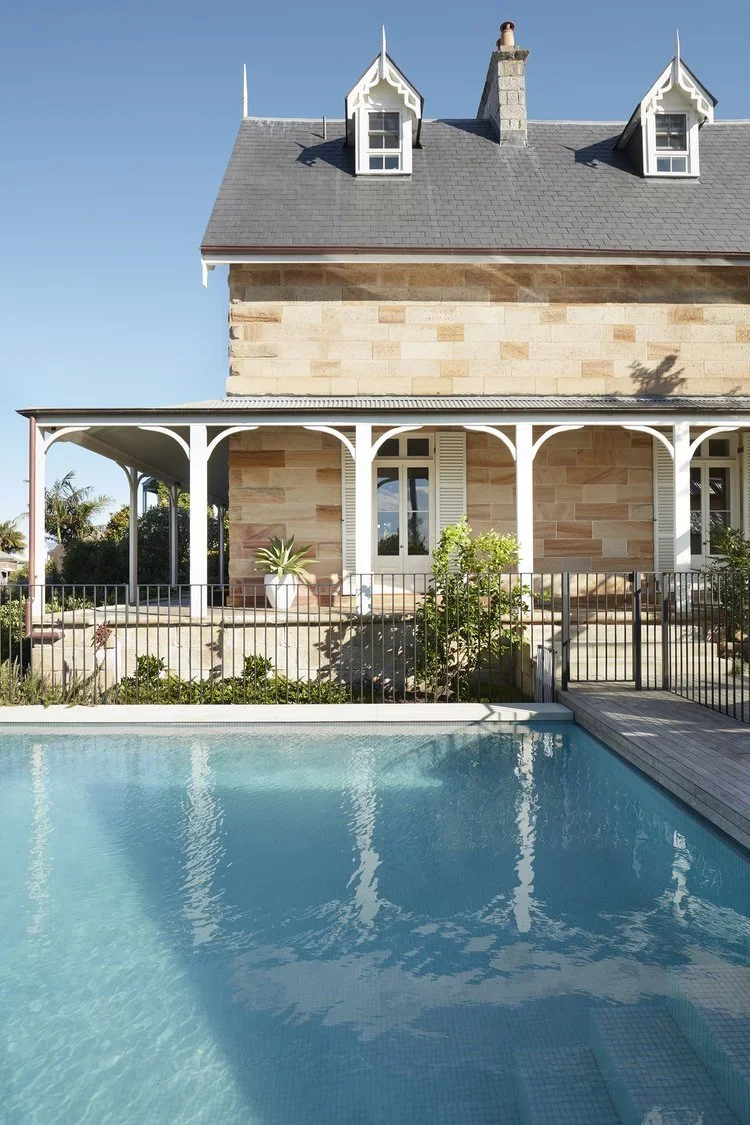 A house with a stone exterior and a covered porch, featuring white trim and a grey roof, alongside a backyard pool with clear blue water.