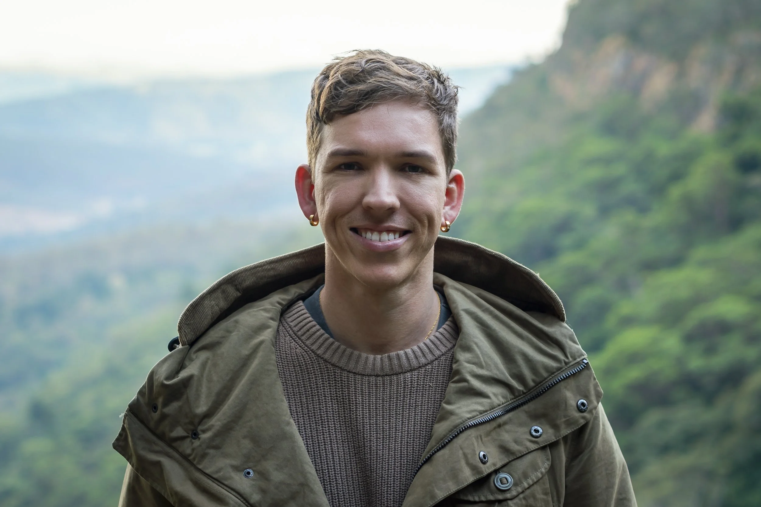 A smiling young woman with short, wavy hair, wearing gold hoop earrings, a brown sweater, and an outdoor jacket, standing in a green, hilly landscape with a cloudy sky in the background.