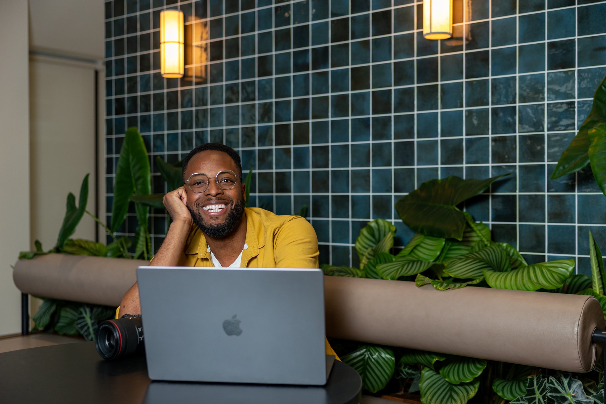 Smiling man in a mustard yellow shirt working at a laptop by a teal tiled wall with green plants | Corporate headshots Victoria, London | Shot By Andrew