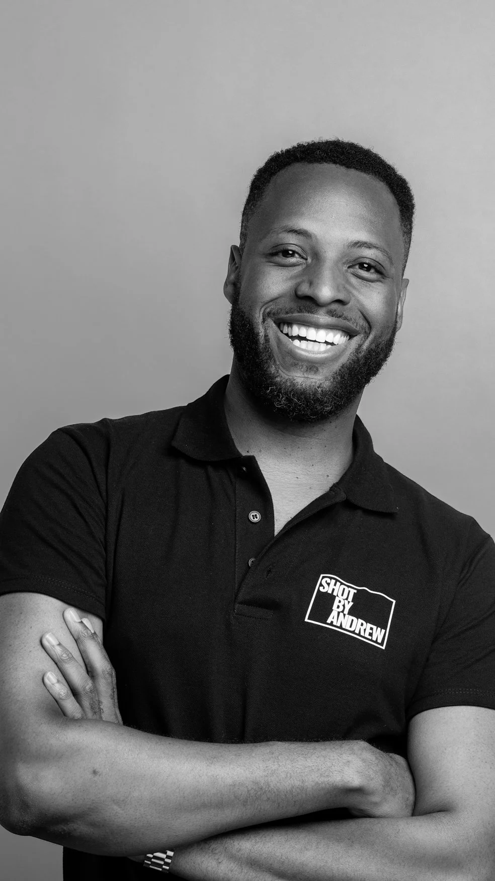 Black and white image of the company headshot photographer smiling. He’s a bearded man with folded arms in a black branded polo | Shot By Andrew