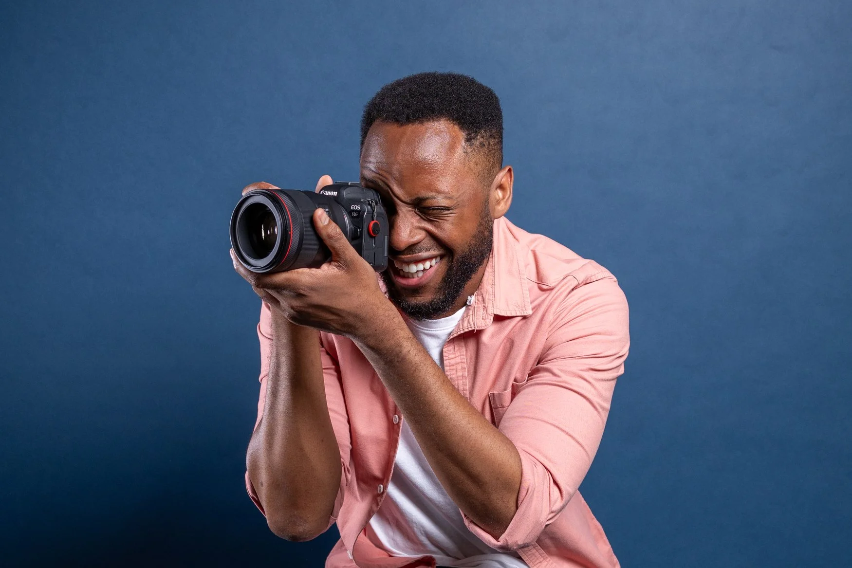 Smiling photographer in a pink shirt shooting with a camera to his eye against a deep blue studio background | Corporate headshots Vauxhall, London | Shot By Andrew