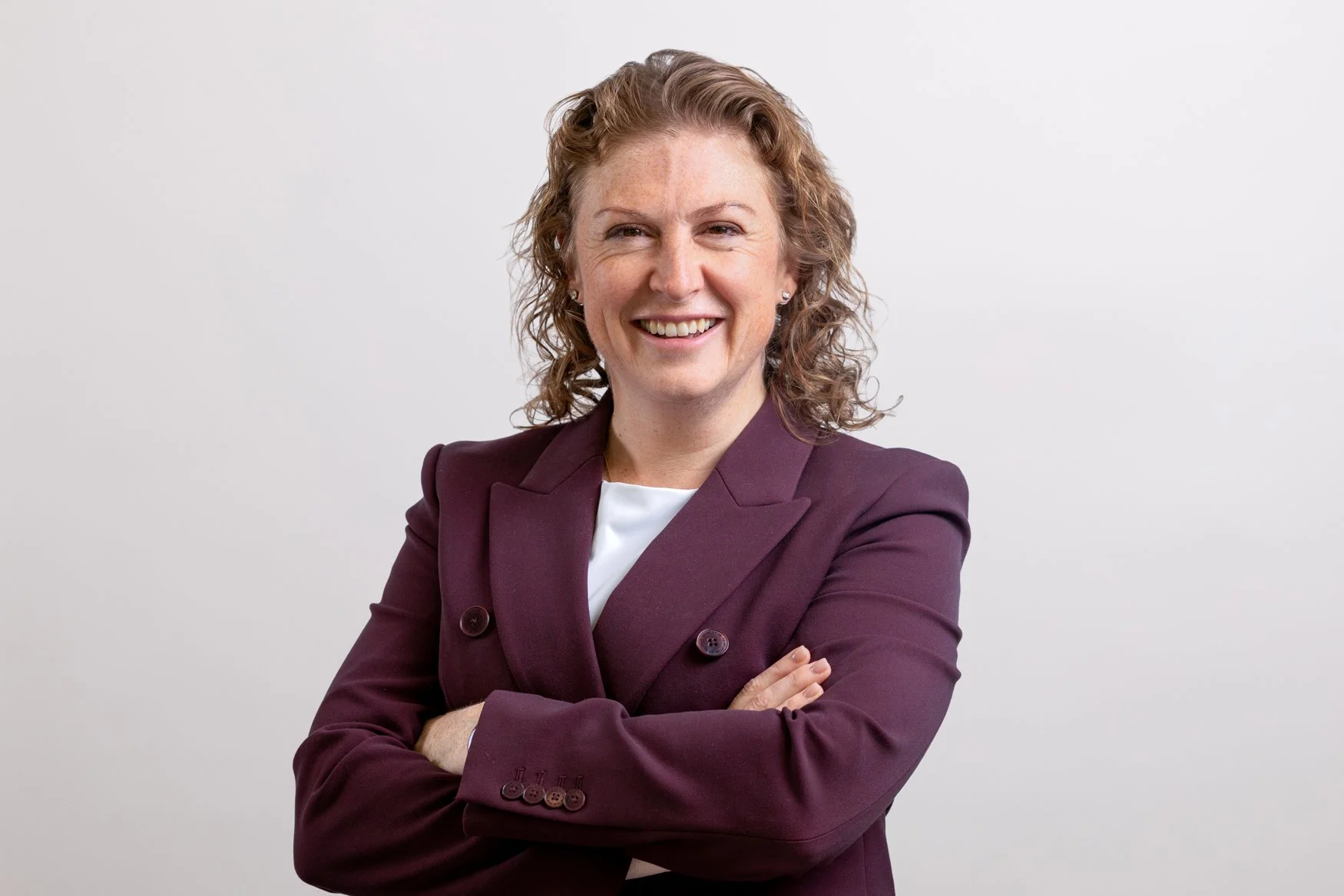 Corporate headshot of a female executive in a burgundy blazer with arms folded on a grey background