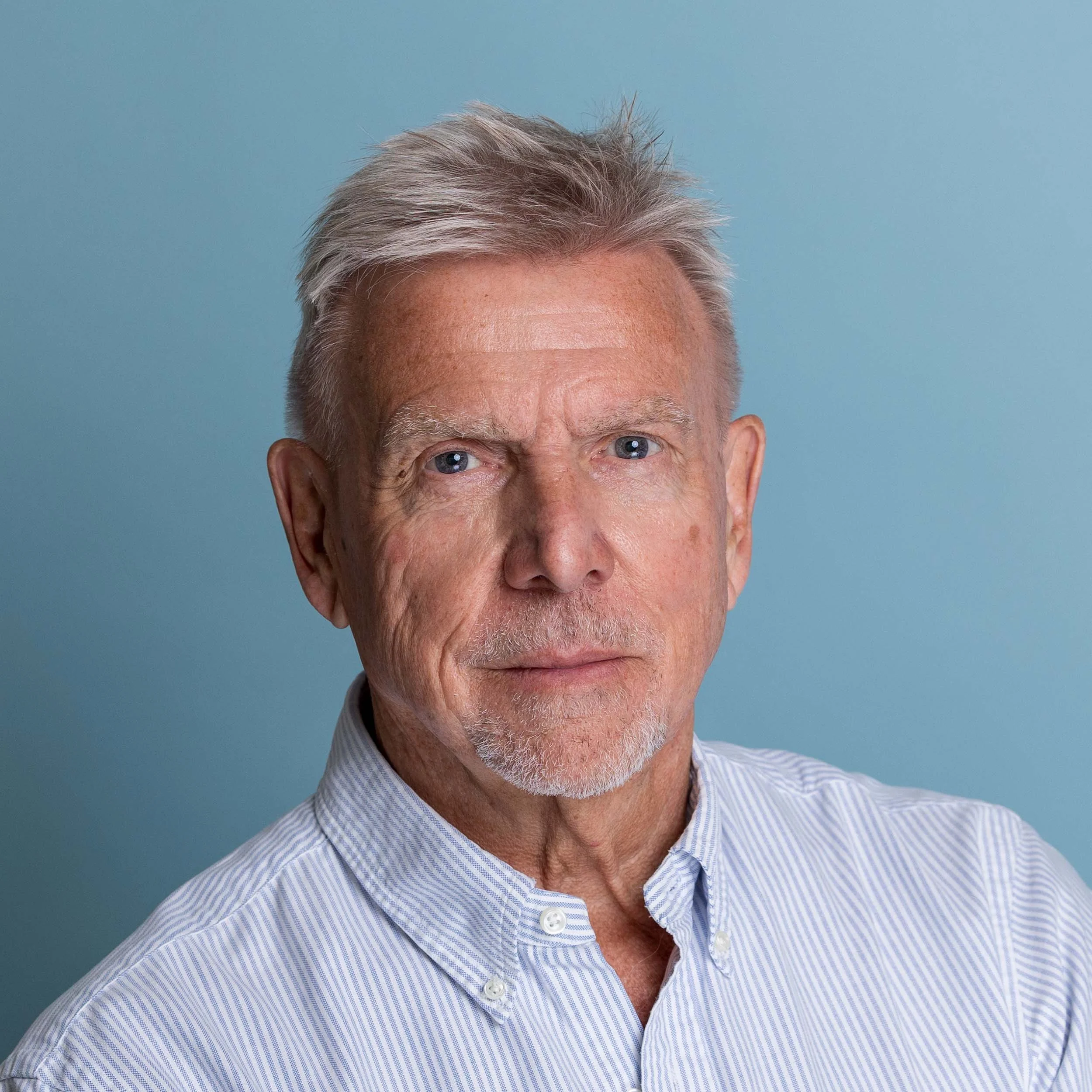 Studio image of an older man with blue eyes looking straight into camera taking part in a linkedin headshot session at the shot by andrew studios. He has a lined purple shirt on