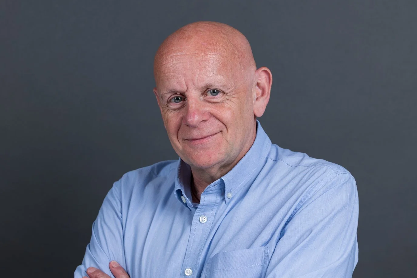 Older bald man in a light blue shirt, arms crossed with a warm smile on a blue-grey background | Business headshots London | Shot By Andrew