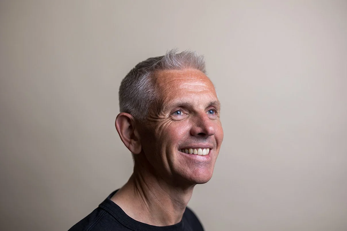 Middle-aged man with grey-flecked hair in a black t-shirt beaming with a natural, relaxed smile — professional LinkedIn profile photos London | Shot By Andrew