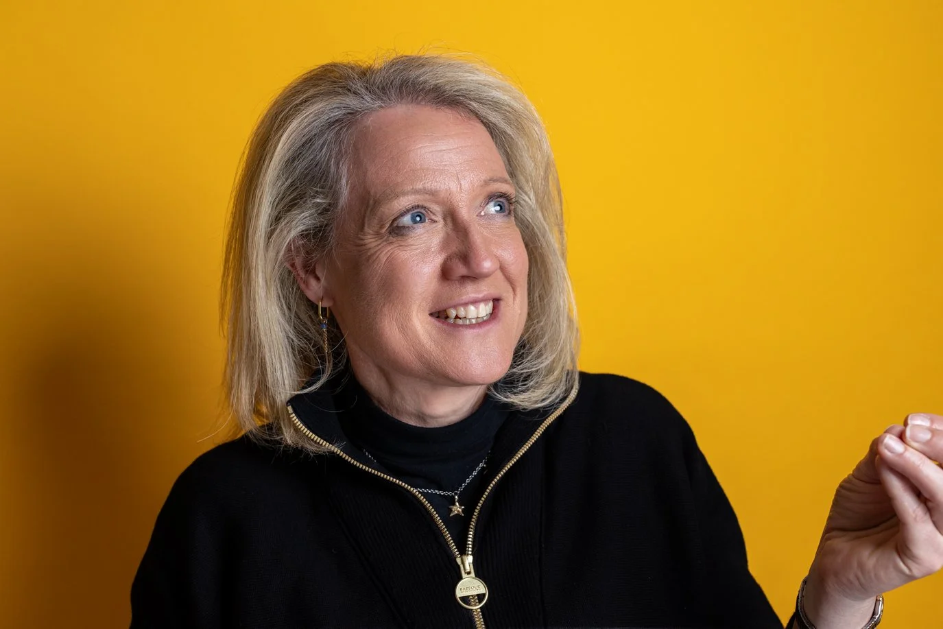 Woman with shoulder-length blonde hair in a black top and gold necklace, looking to the side and smiling on a yellow background | Business headshots London | Shot By Andrew