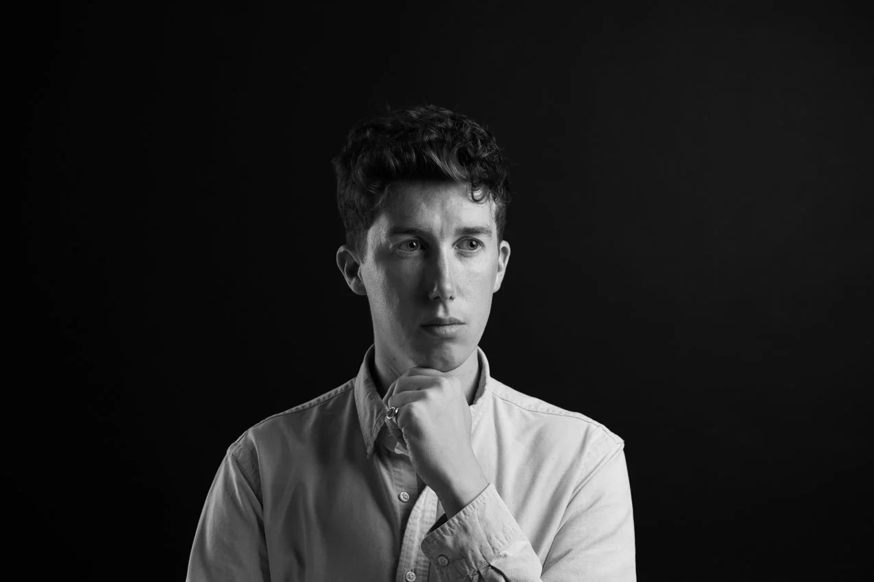 Young man with dark curly hair in a white shirt resting a hand on his chin, thoughtful expression on a black background, black and white | Business headshots London | Shot By Andrew