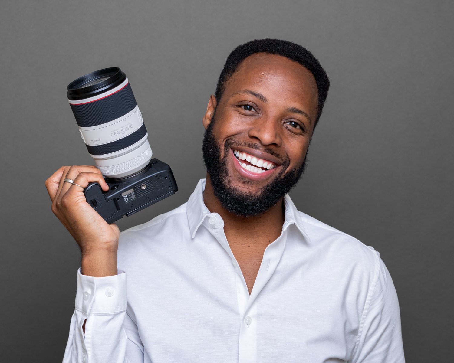 Smiling bearded man in a white shirt holding a camera with a long white lens, dark grey background | Corporate headshots Victoria, London | Shot By Andrew