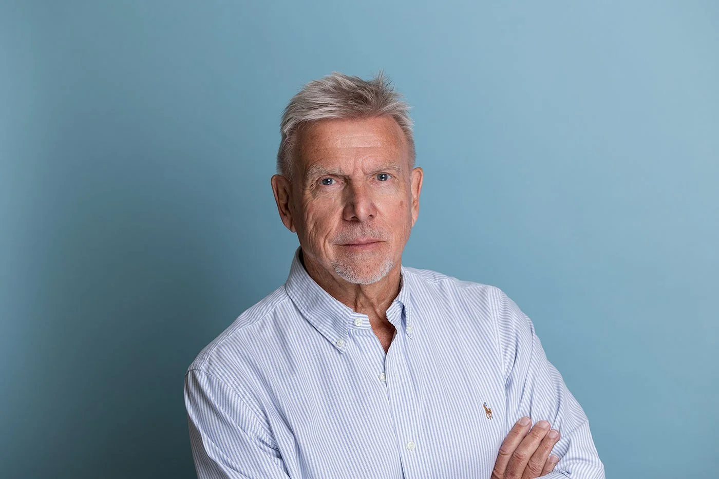 Older white man with silver hair wearing a light blue Ralph Lauren striped polo shirt, arms crossed, against a deep teal studio background | Corporate headshots City of London | Shot By Andrew
