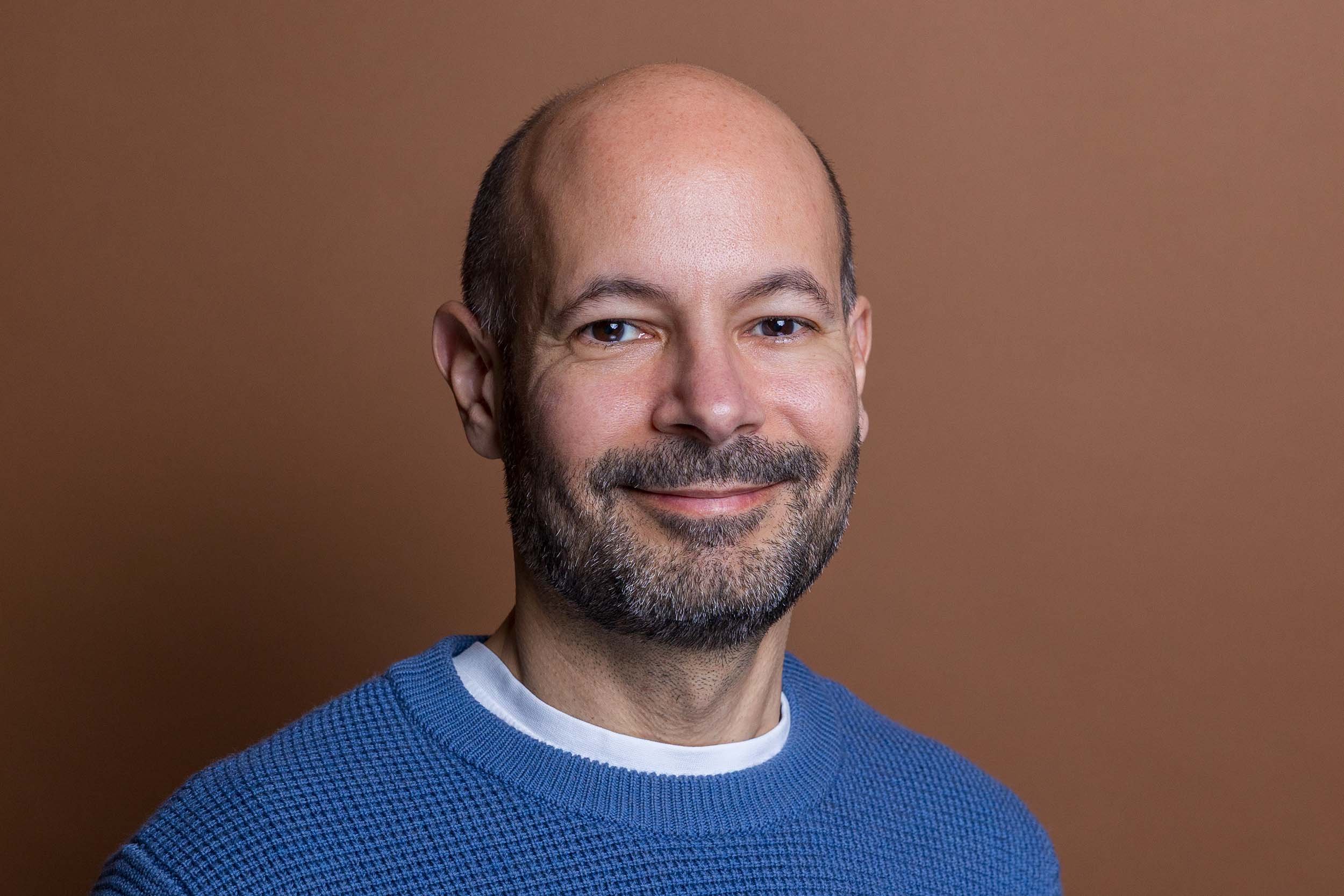 Studio image of a man with brown eyes looking straight into camera taking place in a linkedin headshot session at the shot by andrew studios. He has a blue jumper on with a white t-shirt underneath