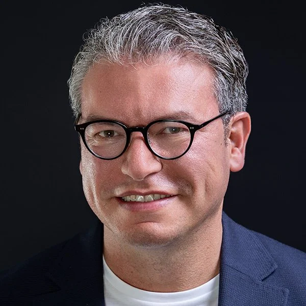 Man with salt-and-pepper hair and thin-framed glasses in a dark jacket, warm approachable smile — LinkedIn headshots London | Shot By Andrew