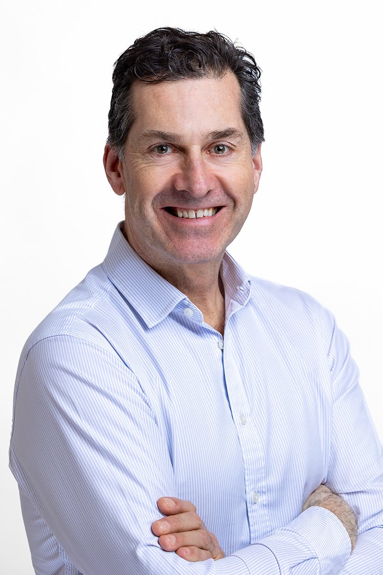 Middle-aged professional man with dark wavy hair, arms crossed and smiling broadly, in a light blue striped shirt against a white background — Mayfair headshot | Shot By Andrew