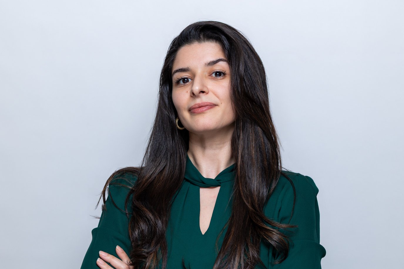 Woman with long dark hair in a dark green blouse, arms crossed with a subtle smile on a pale grey background | Business headshots London | Shot By Andrew