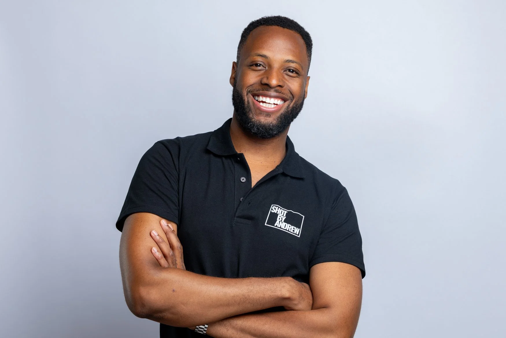 London Corporate Photographer standing in a black branded polo shirt, smiling with arms crossed.