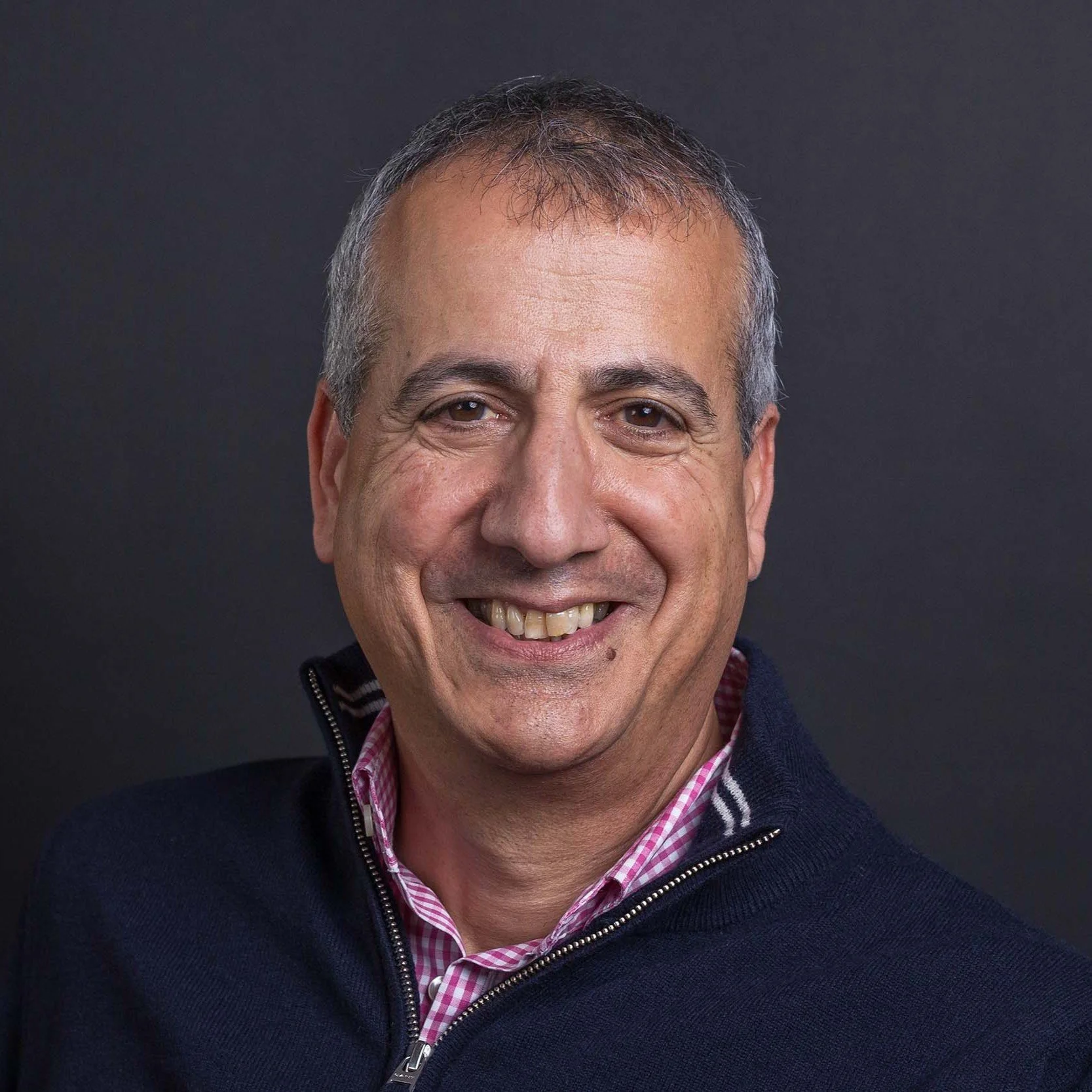 Older professional man with silver curly hair in a navy vest over a pink check shirt, broad warm smile, dark grey background — Mayfair headshot | Shot By Andrew