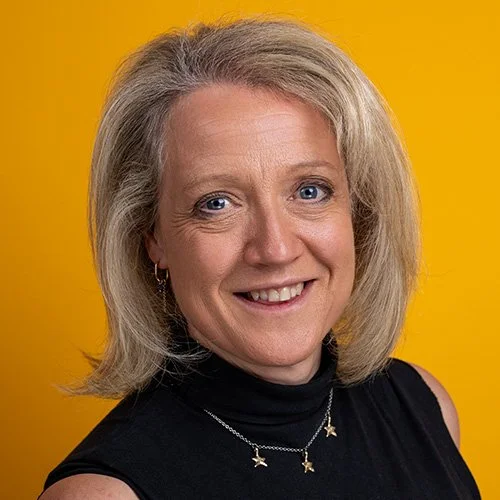 Blonde woman in a black turtleneck with gold star necklace, warm smile against a bright yellow background — London LinkedIn headshot example | Shot By Andrew