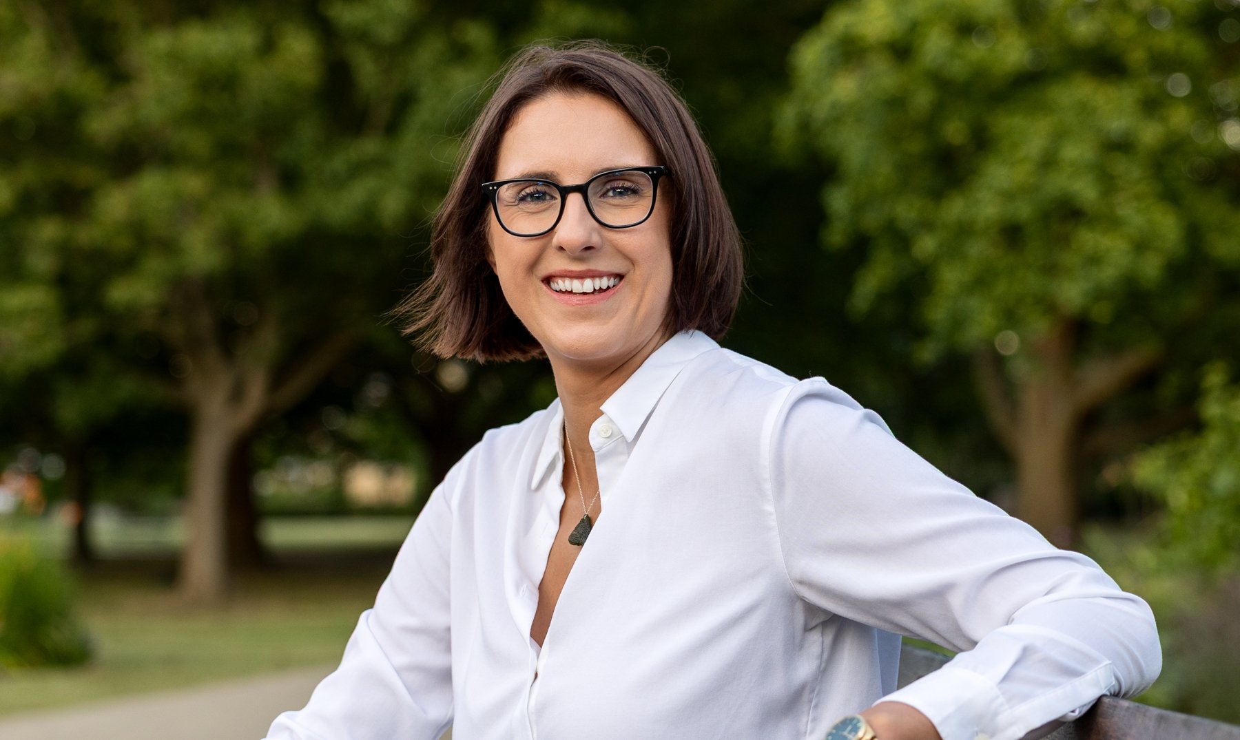 Outdoor headshot of a smiling woman in glasses and a white shirt, seated in a park with trees behind her, by Shot By Andrew