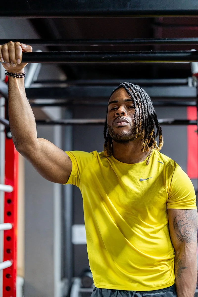 Fitness professional with braided hair in a yellow t-shirt, arm raised at a gym bar — illustrating industry-appropriate LinkedIn profile photo attire | Shot By Andrew London
