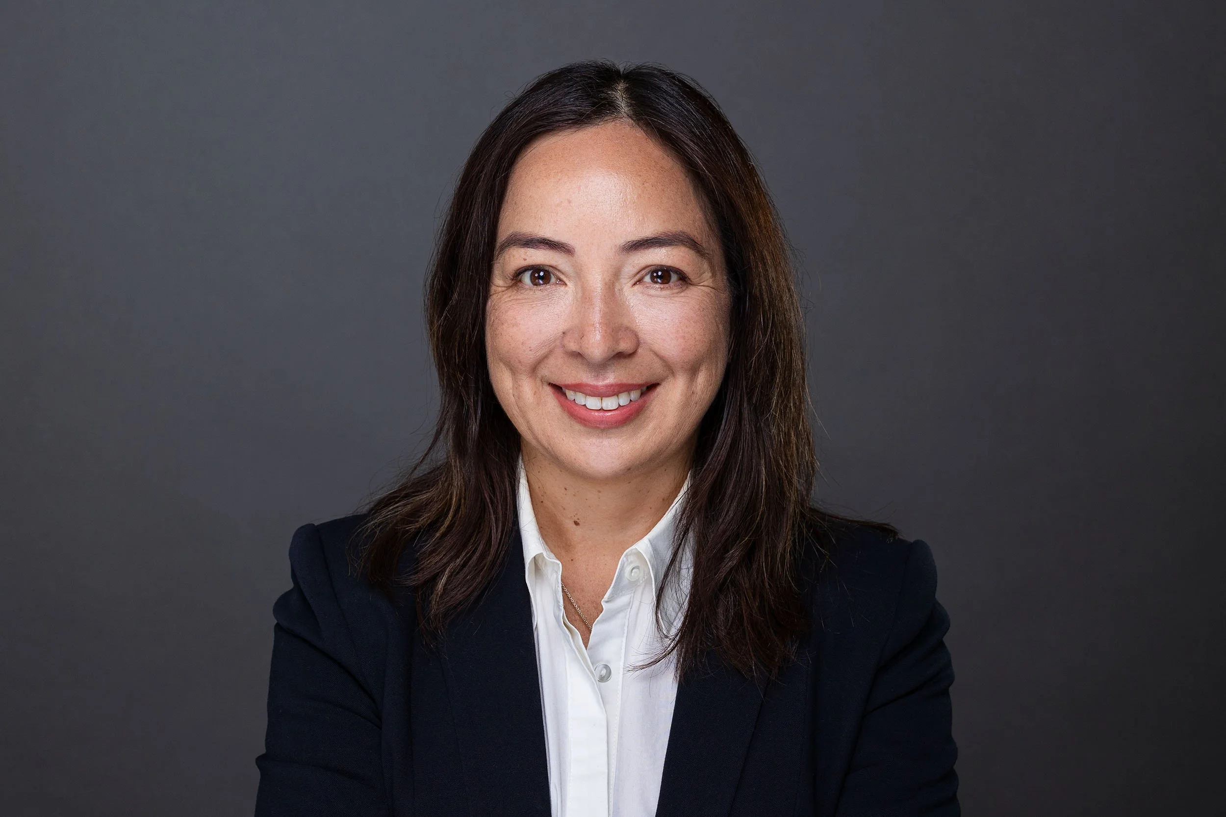 Studio image of a woman looking straight on and right into camera taking place in a linkedin headshot session at the shot by andrew studios. she is in a white shirt with a black blazer