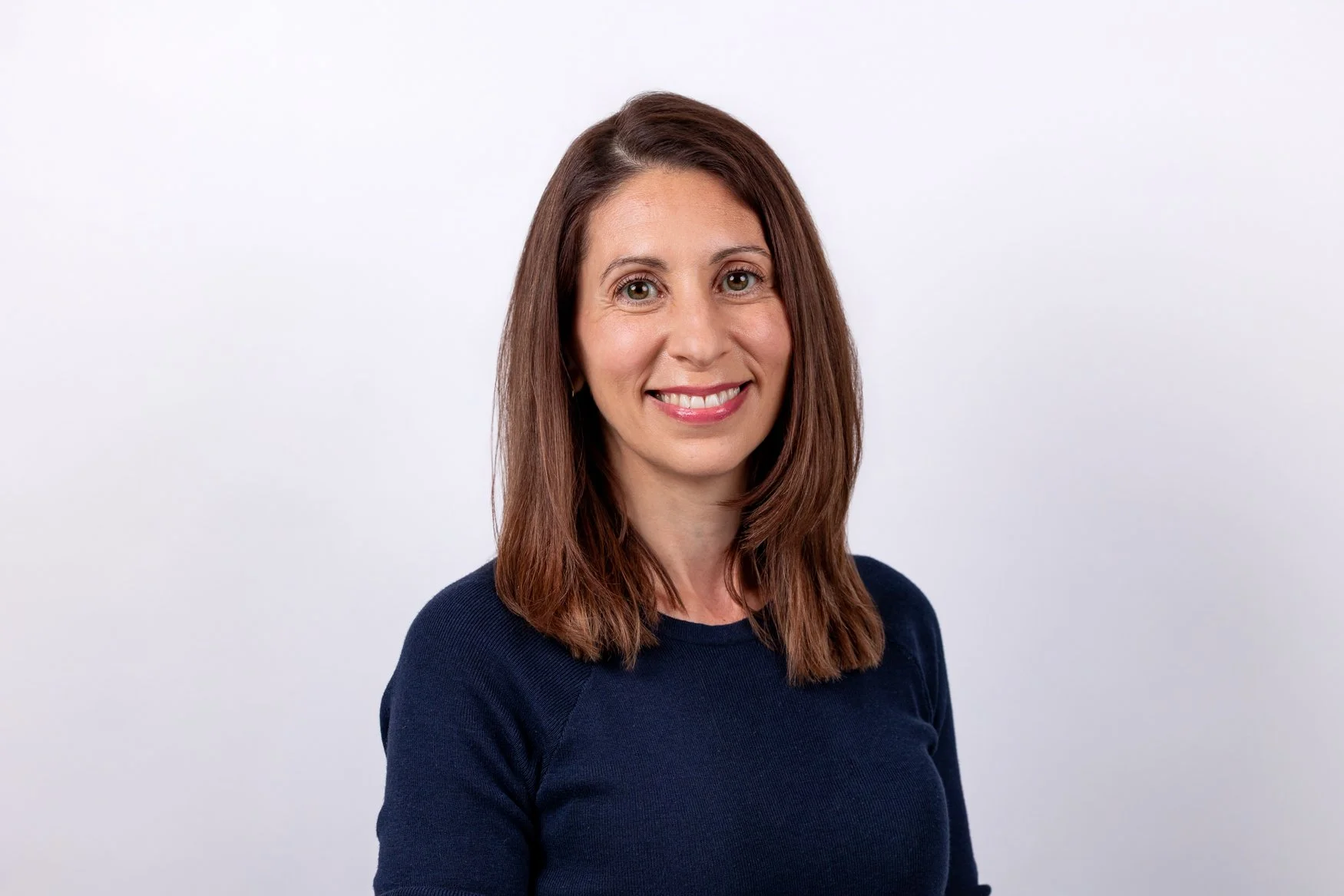 Corporate headshot of a smiling woman with long auburn hair in a navy top, soft white-grey background | London headshot photographer | Shot By Andrew