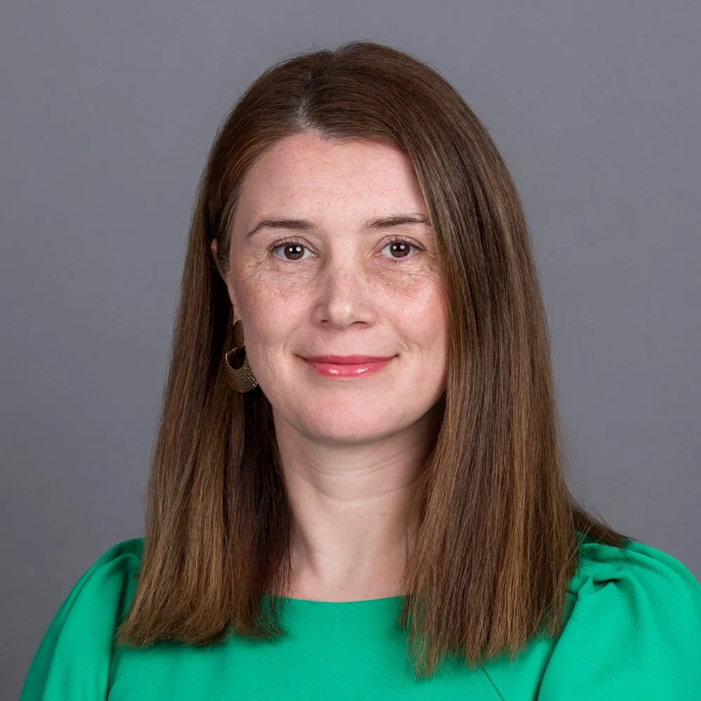 Corporate headshot of a woman in green with mid length brown hair against a grey backdrop, photographed on location at a data management company in Waterloo, London