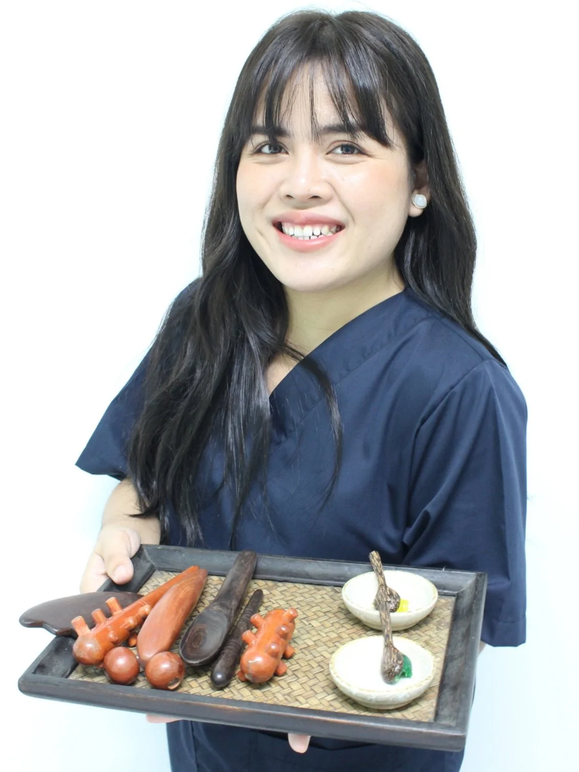 Kvinde i blå uniform, der smiler og holder en træbakke med legetøj, herunder nogle træknive og forskellige små skåle med skeer.