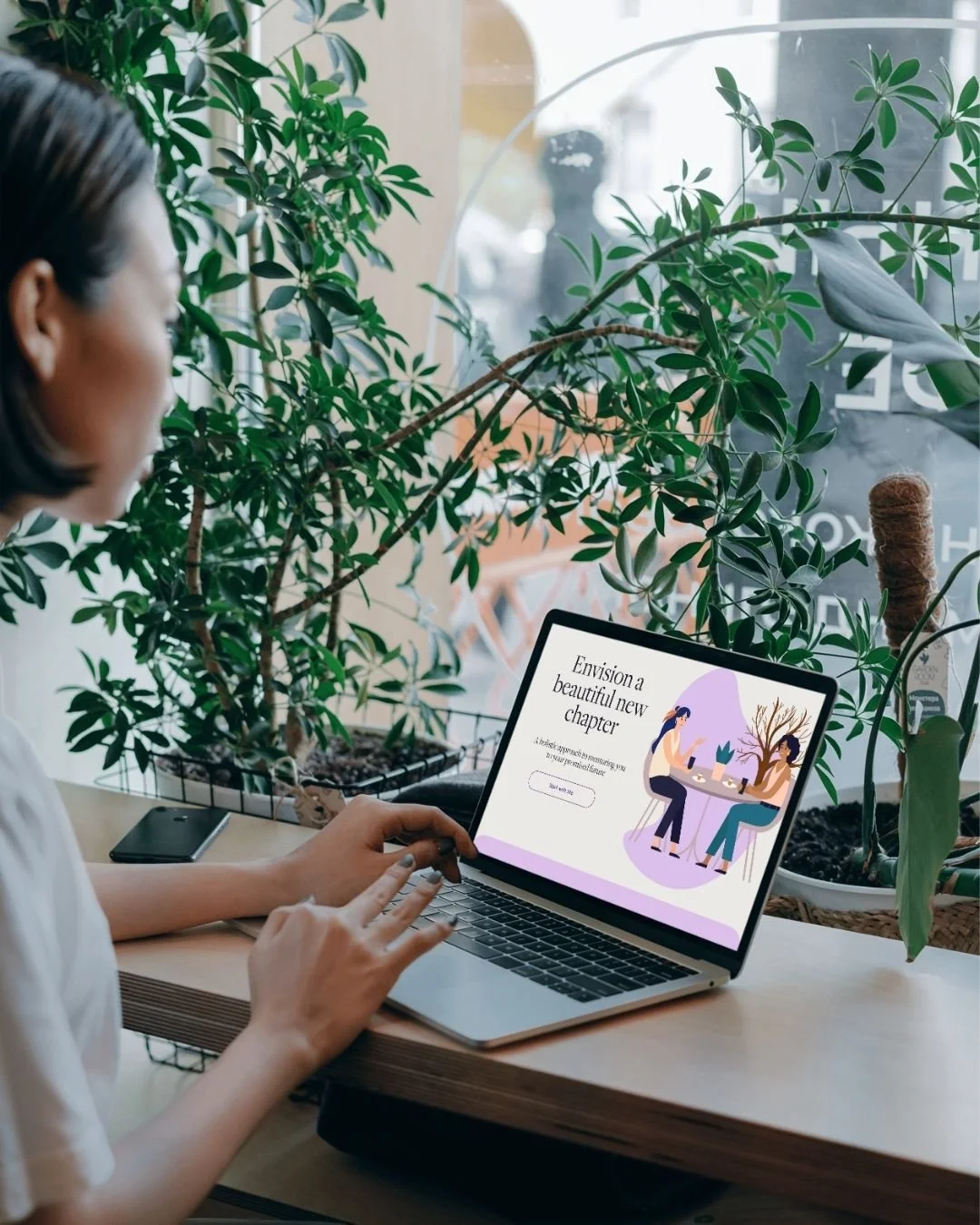 A woman sitting at a wooden table near a window with large green houseplants, looking at a laptop screen displaying a graphic with two women, one holding a phone and the other cute trees, and the text 'Envision a beautiful new chapter'.
