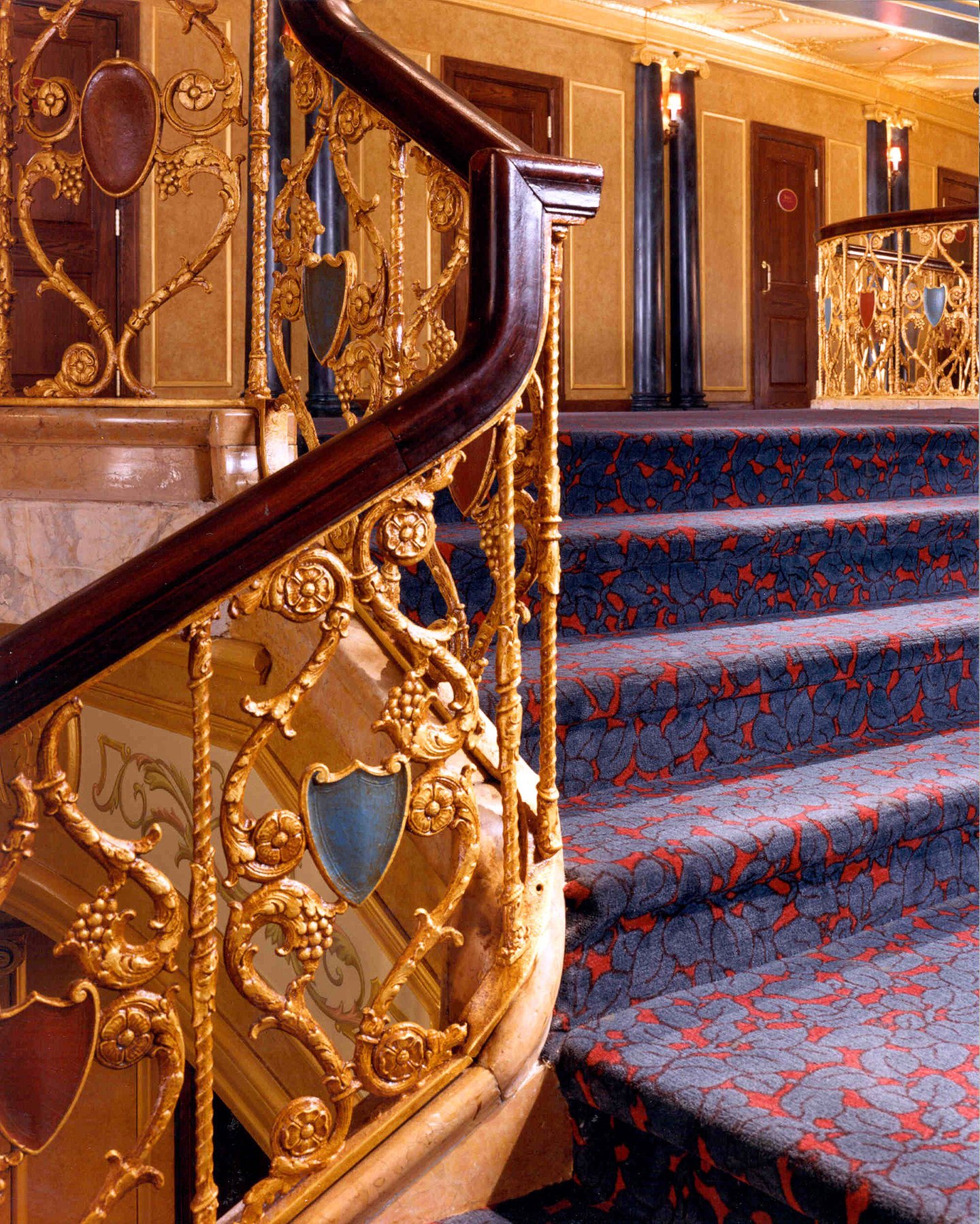 Luxury staircase with an ornate gold railing and a dark wood handrail, carpeted steps with a floral pattern in red and blue, elegant gold-paneled walls.
