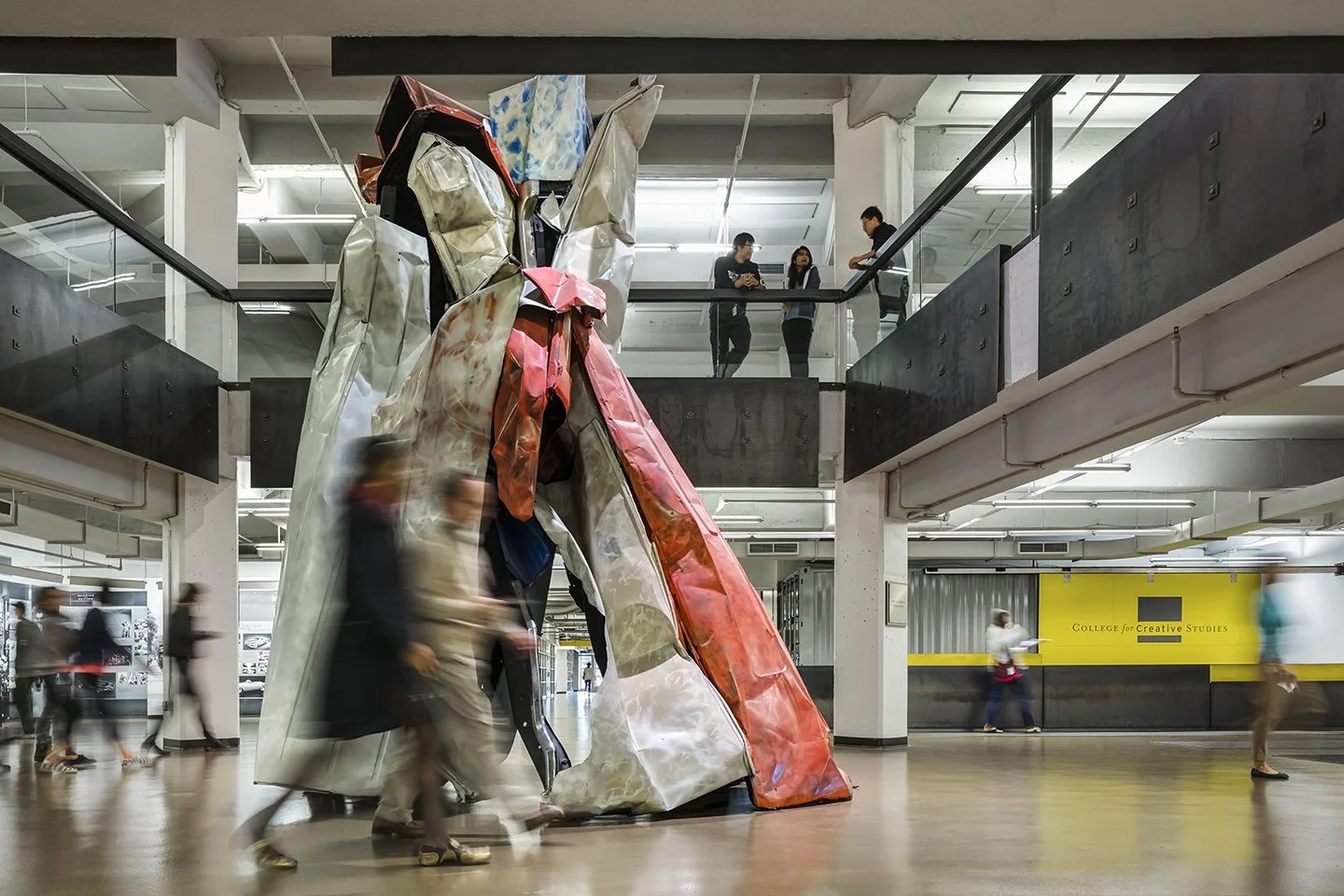 A large contemporary art sculpture made of crumpled metallic and colored materials is displayed in an indoor public space with many walking people. Two individuals are standing on a balcony above, observing the scene.